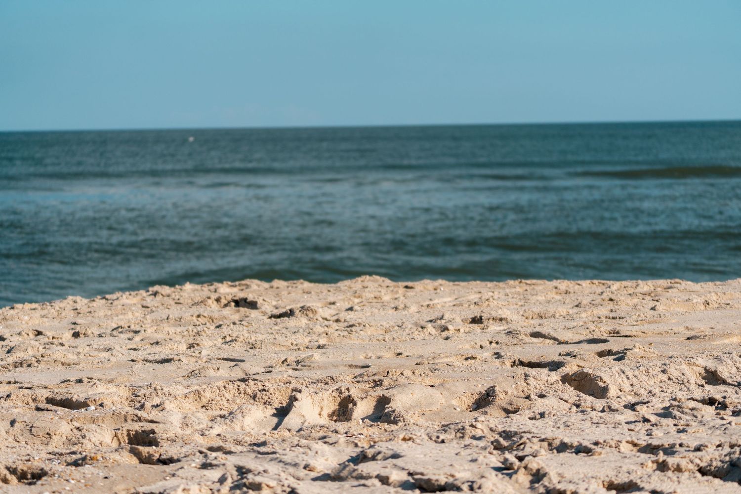 A close-up of a sandy beach with small rocks and the blue ocean horizon in the background.