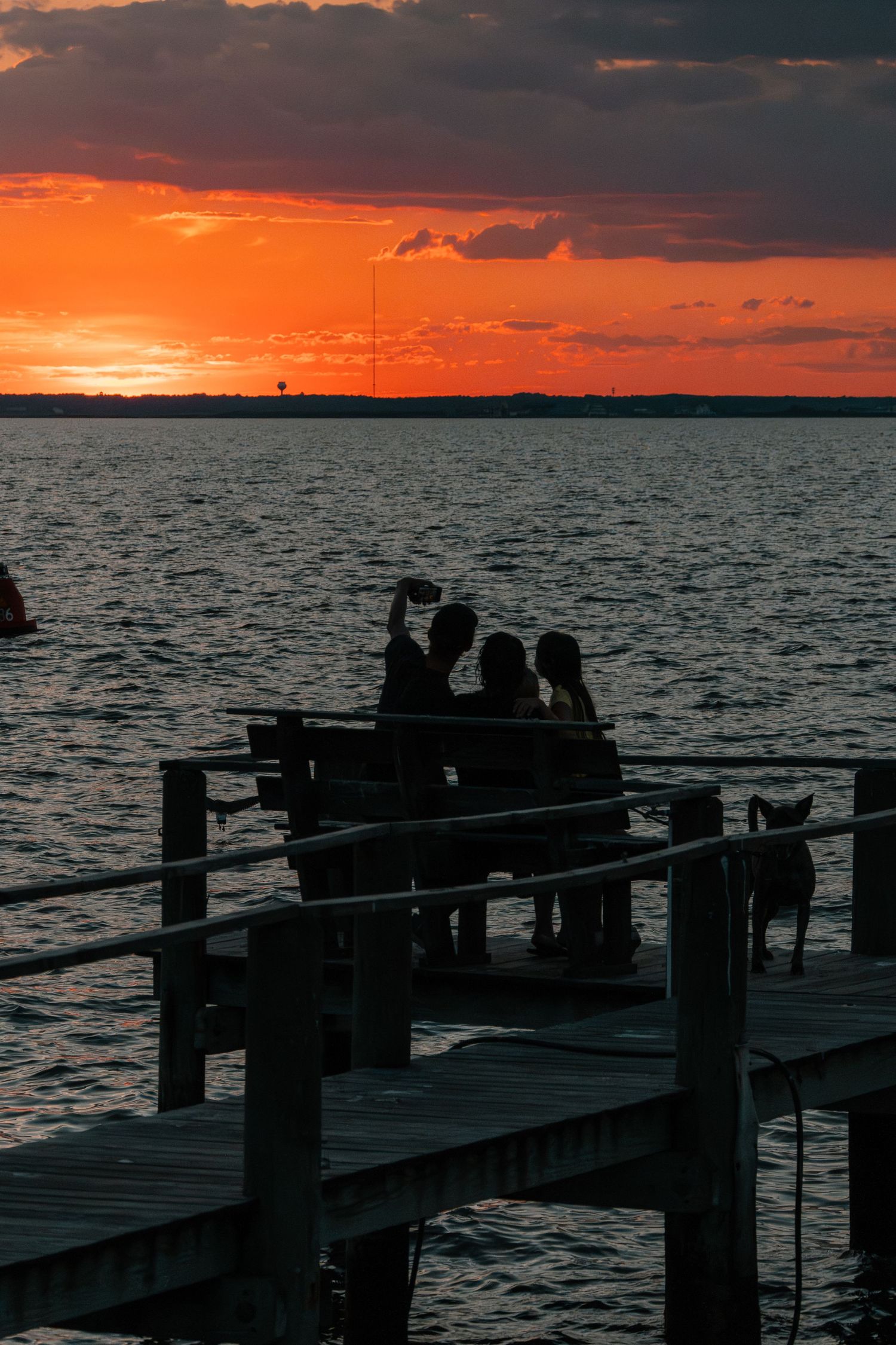 Silhouettes of people on a wooden dock taking a selfie against a vibrant orange sunset over calm water.
