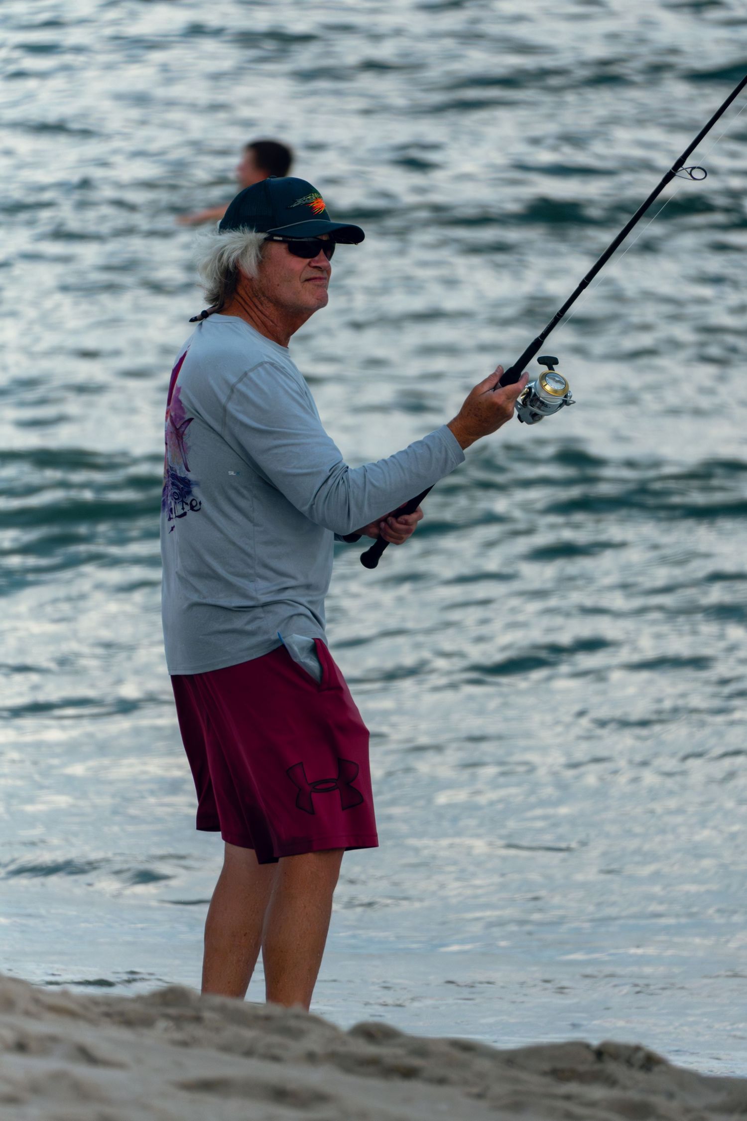 Smiling woman in cap and sunglasses surf fishing on a beach at dusk with ocean behind.