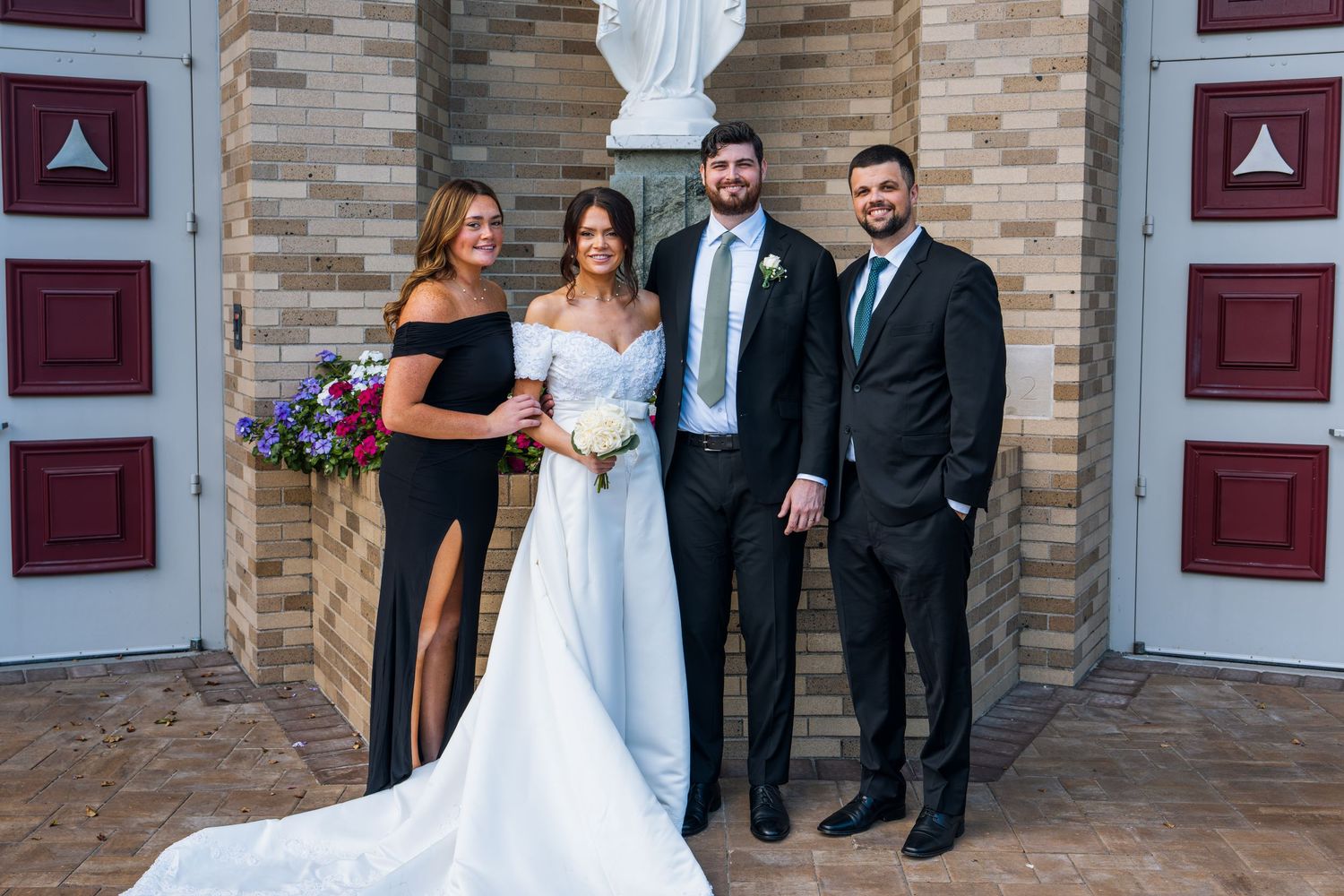 Wedding party of four posing outside a brick church, bride in white gown with groom and guests in formal attire.