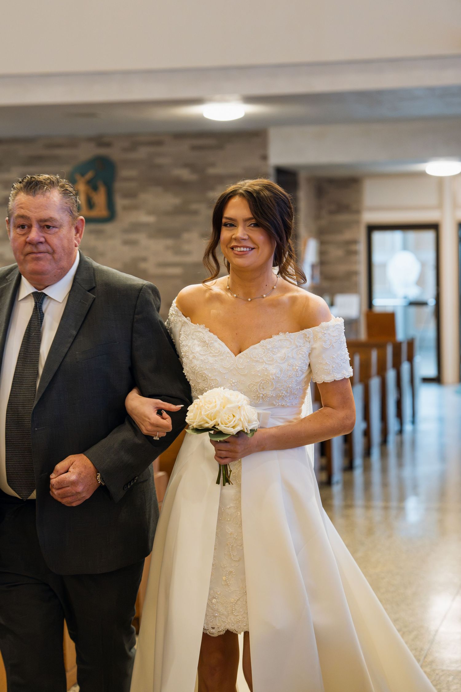 Smiling bride in off-shoulder white gown holding bouquet walks down church aisle with older man in dark suit.