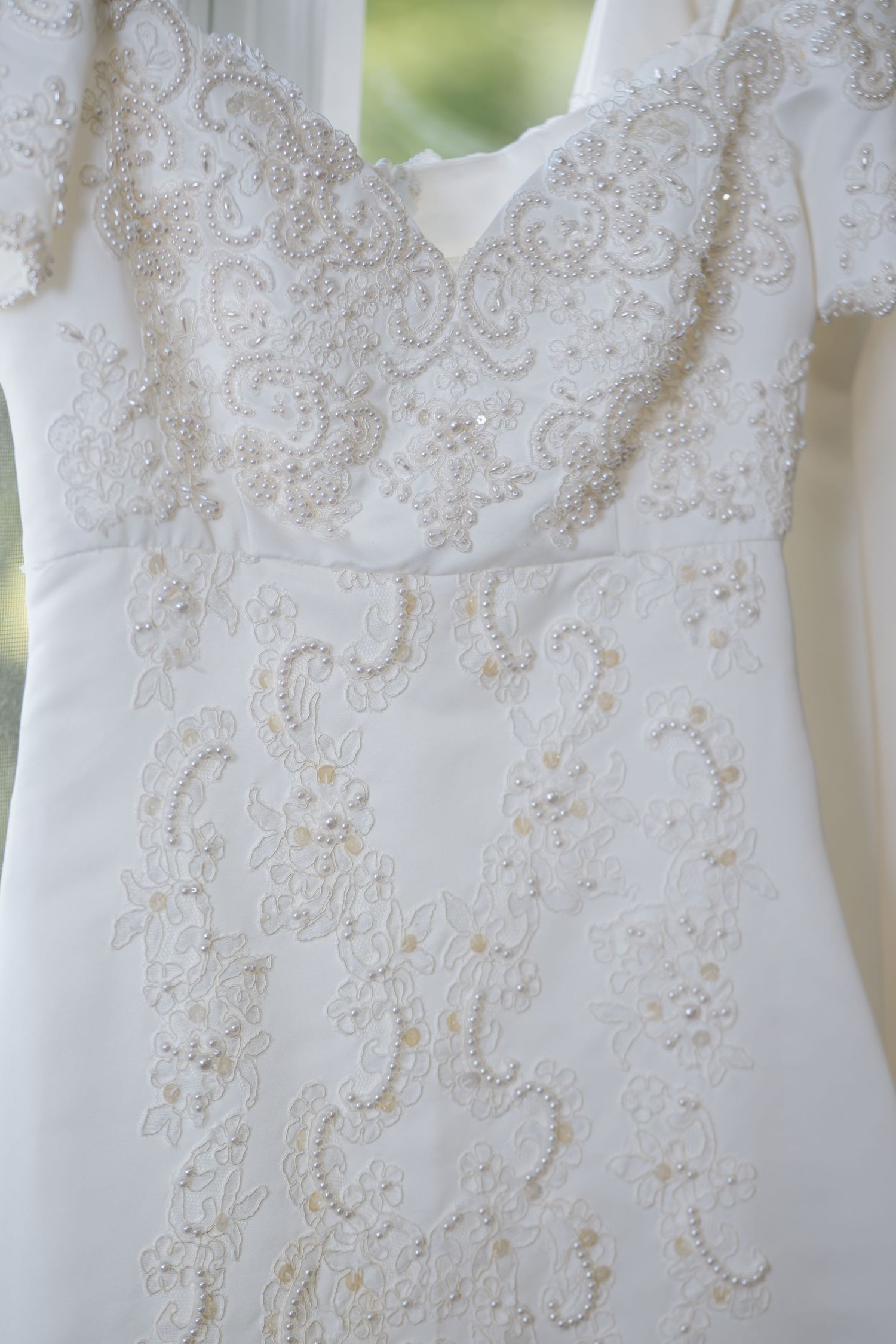 White beaded lace wedding dress with intricate floral embroidery and cap sleeves hanging by a window.