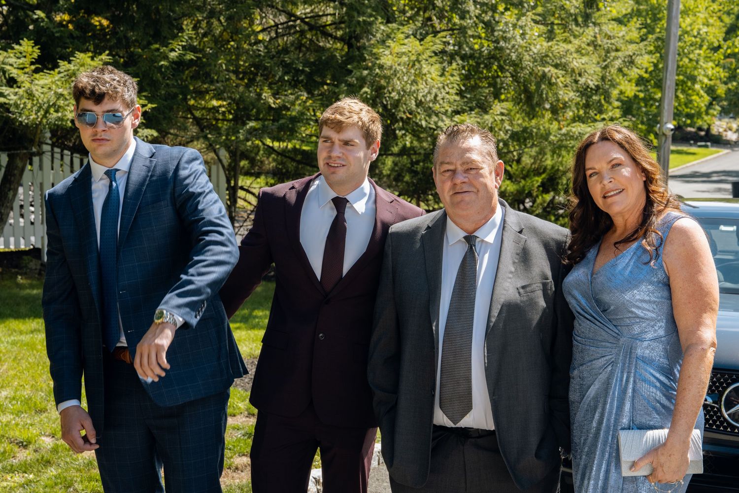 Four formally dressed people posing together outdoors at a sunny event, wearing suits and an elegant blue dress.