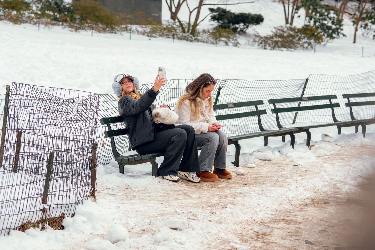 Two women sitting on a snowy park bench, one taking a selfie in a winter landscape.