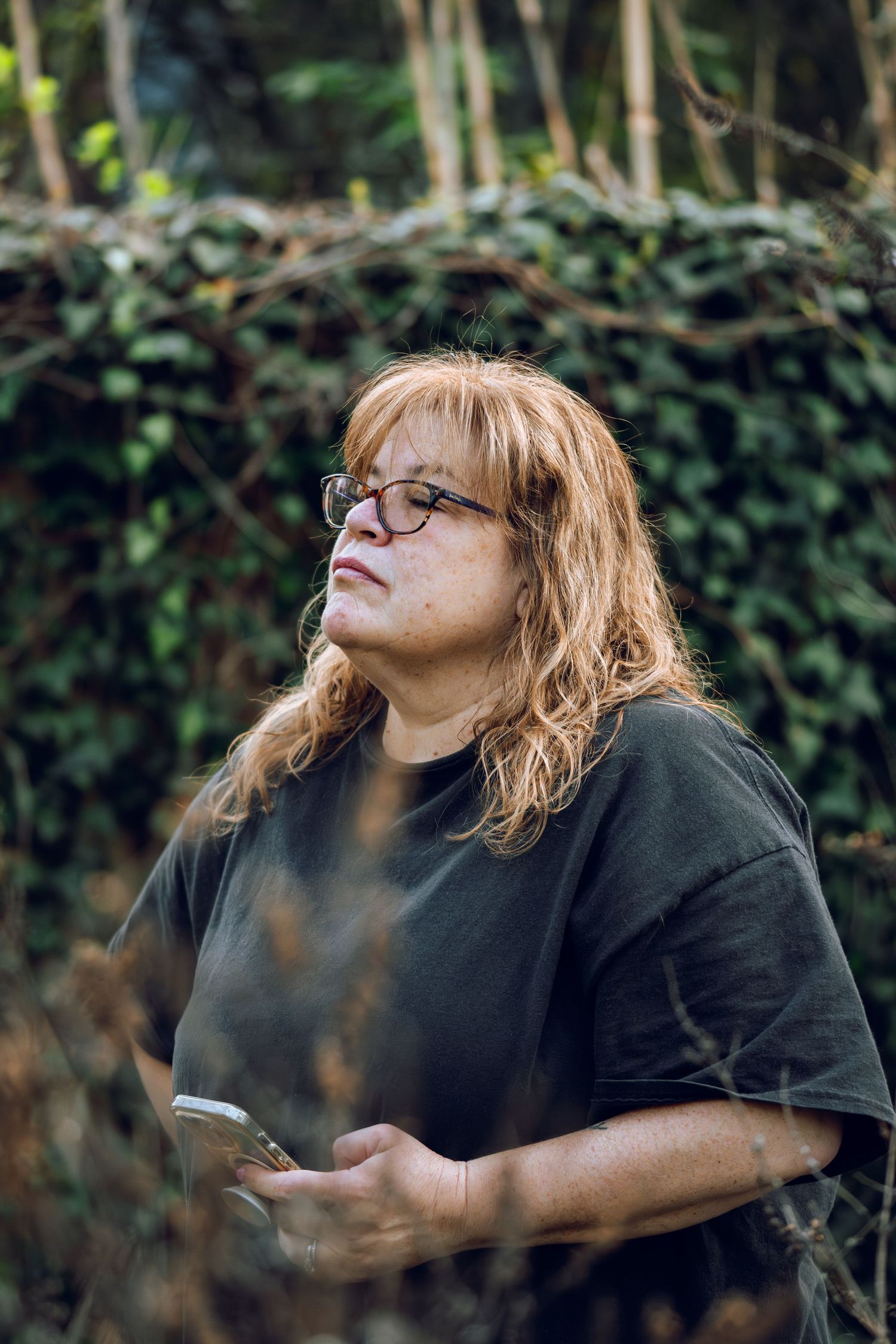 Middle-aged woman with red hair and glasses holding phone outdoors near ivy wall.
