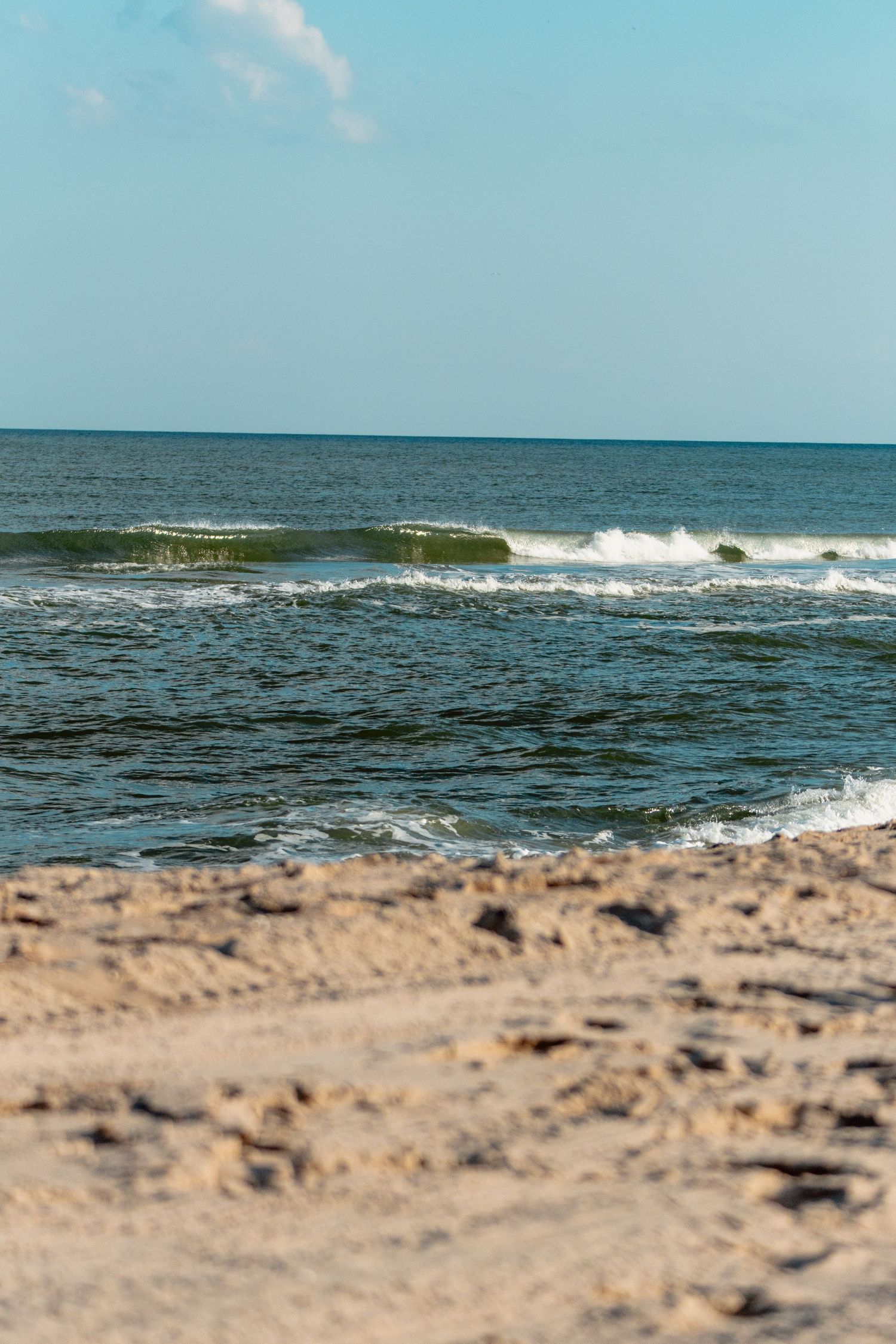 Rolling green ocean waves break gently onto a sandy beach under a bright blue sky on a sunny day.