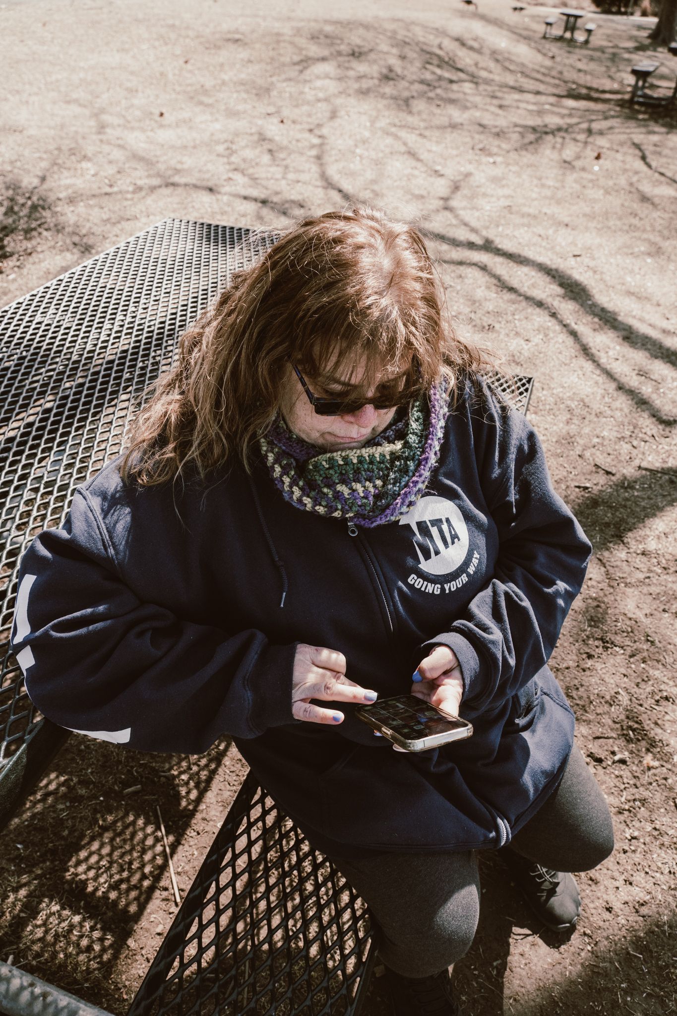 Woman with long hair wearing hoodie and scarf using smartphone on metal bench outdoors.