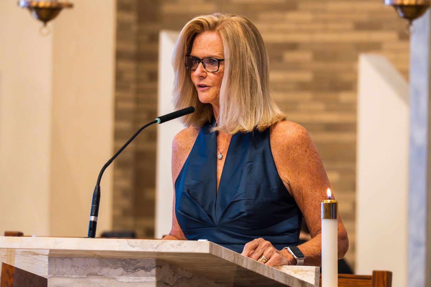 Woman in blue dress speaking at a podium with microphone in a church setting with candle.