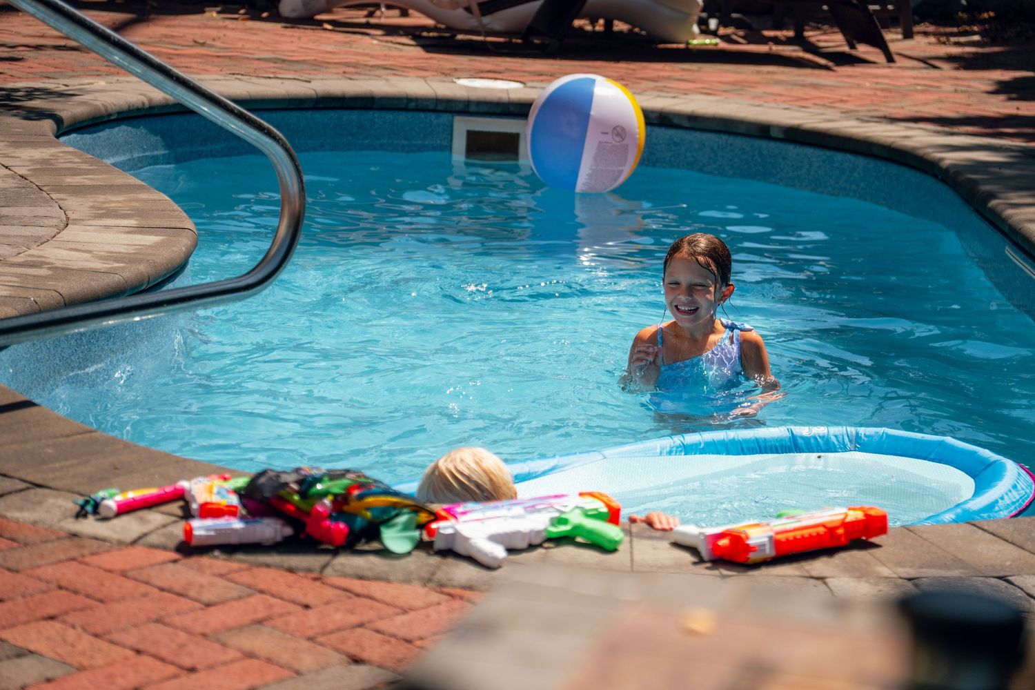 Child swimming in a backyard pool with a beach ball and water toys on the pool deck.