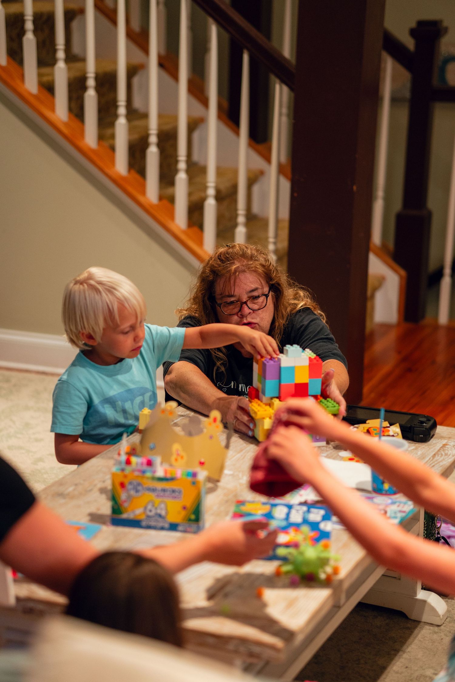 Grandmother helping young blonde boy build colorful blocks at a coffee table during family gathering.