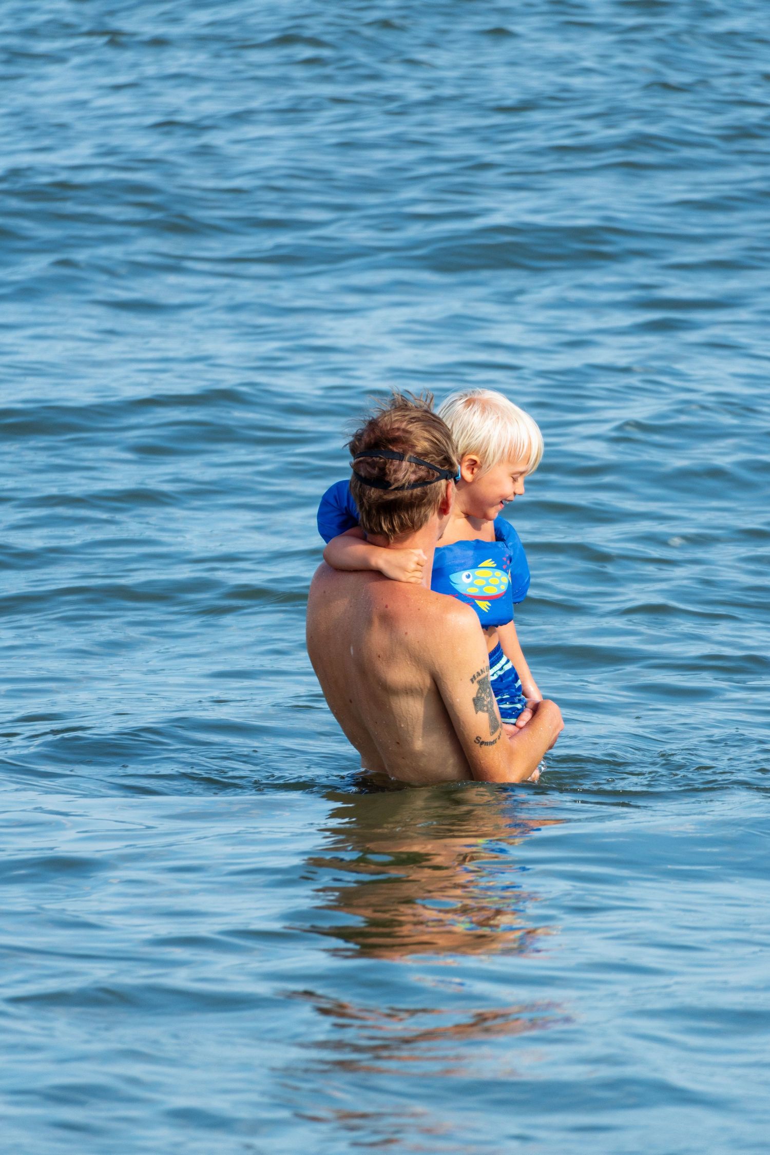 Adult holding a young child wearing a blue life vest while standing in calm ocean water.