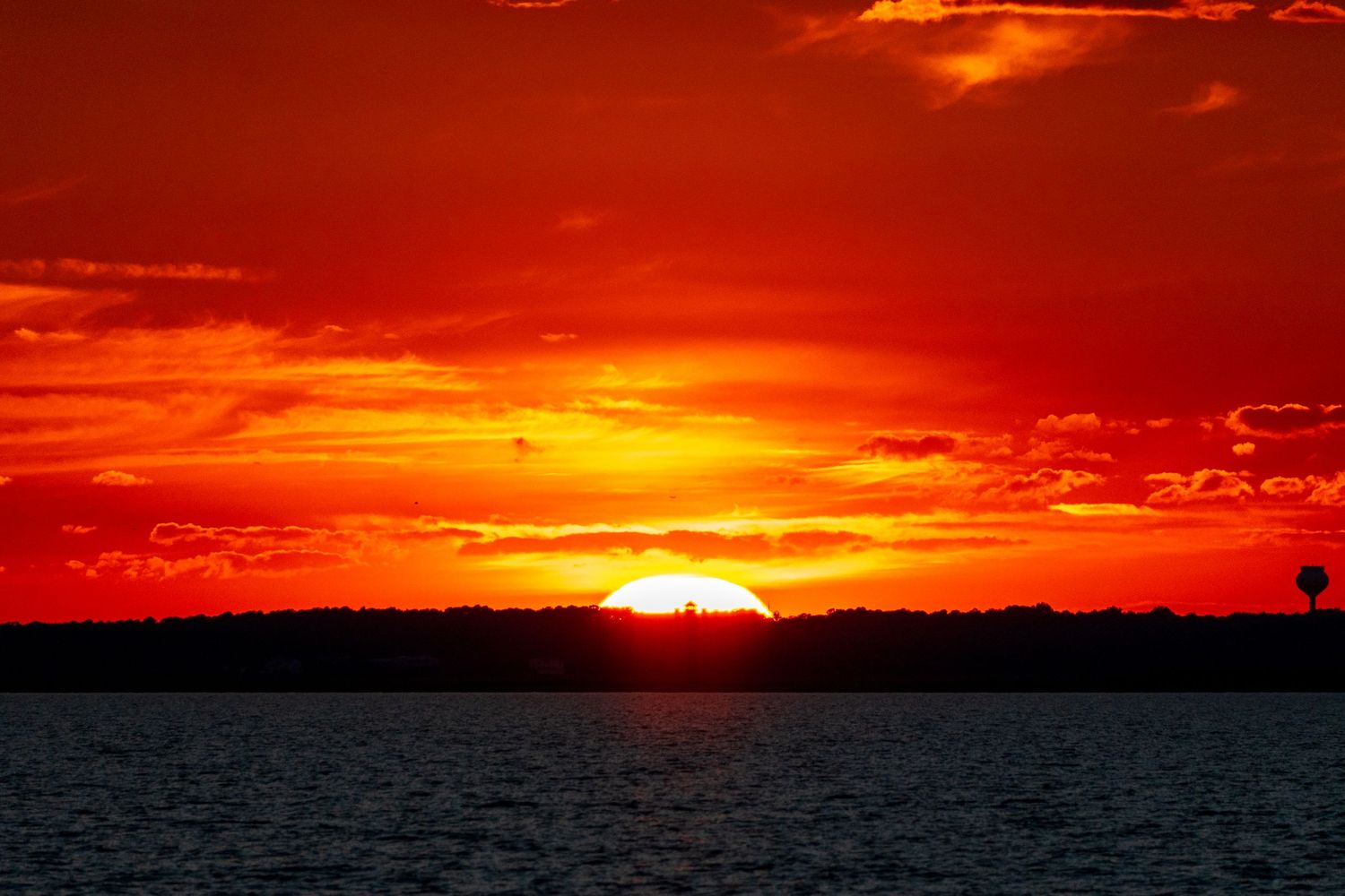 Intense orange and red sunset sinking behind a dark treeline over calm bay waters.