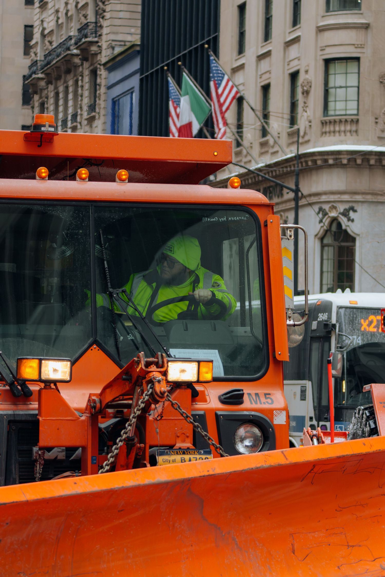 Orange NYC snowplow truck with driver in green jacket clearing city streets in winter.