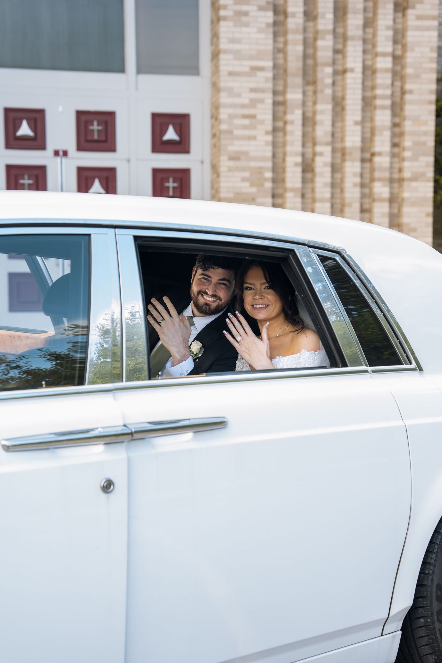 Newlywed couple waving wedding rings from white limousine outside church after ceremony.