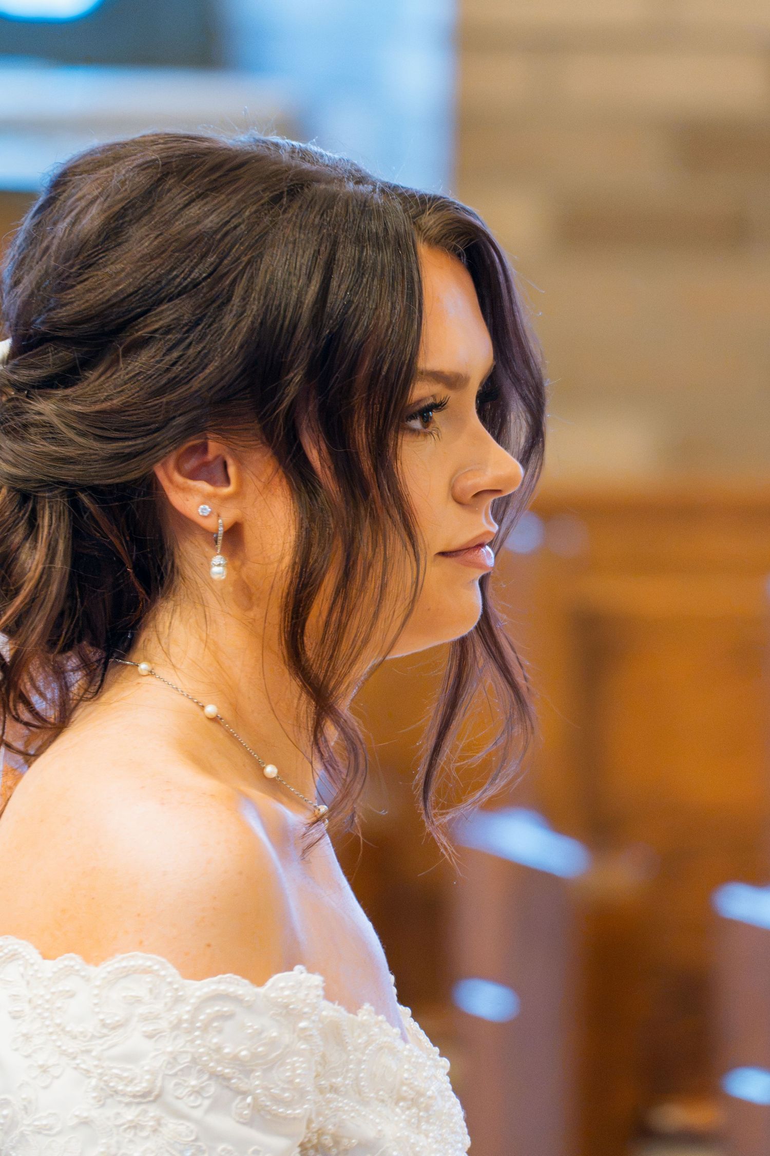 Bride in white off-shoulder lace gown with pearl earrings and necklace, elegant updo hairstyle, profile view in church.