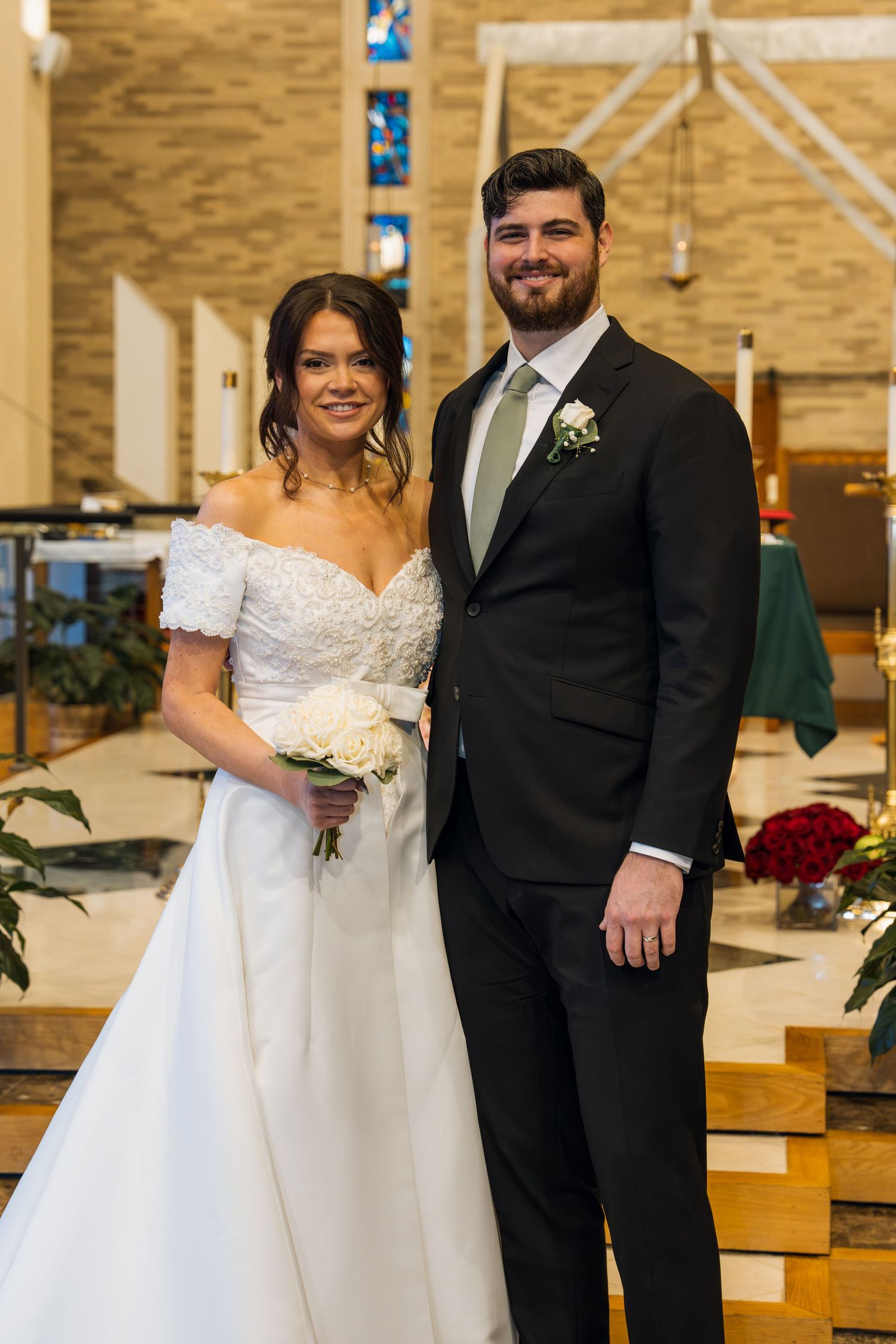 Bride in white off-shoulder gown holding bouquet stands with groom in black suit and green tie inside a church.