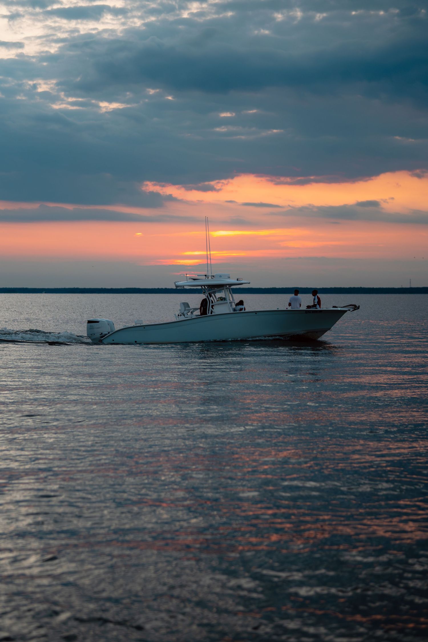 A white motorboat cruises across calm waters at sunset with a sailboat silhouetted against a vibrant orange and pink sky.