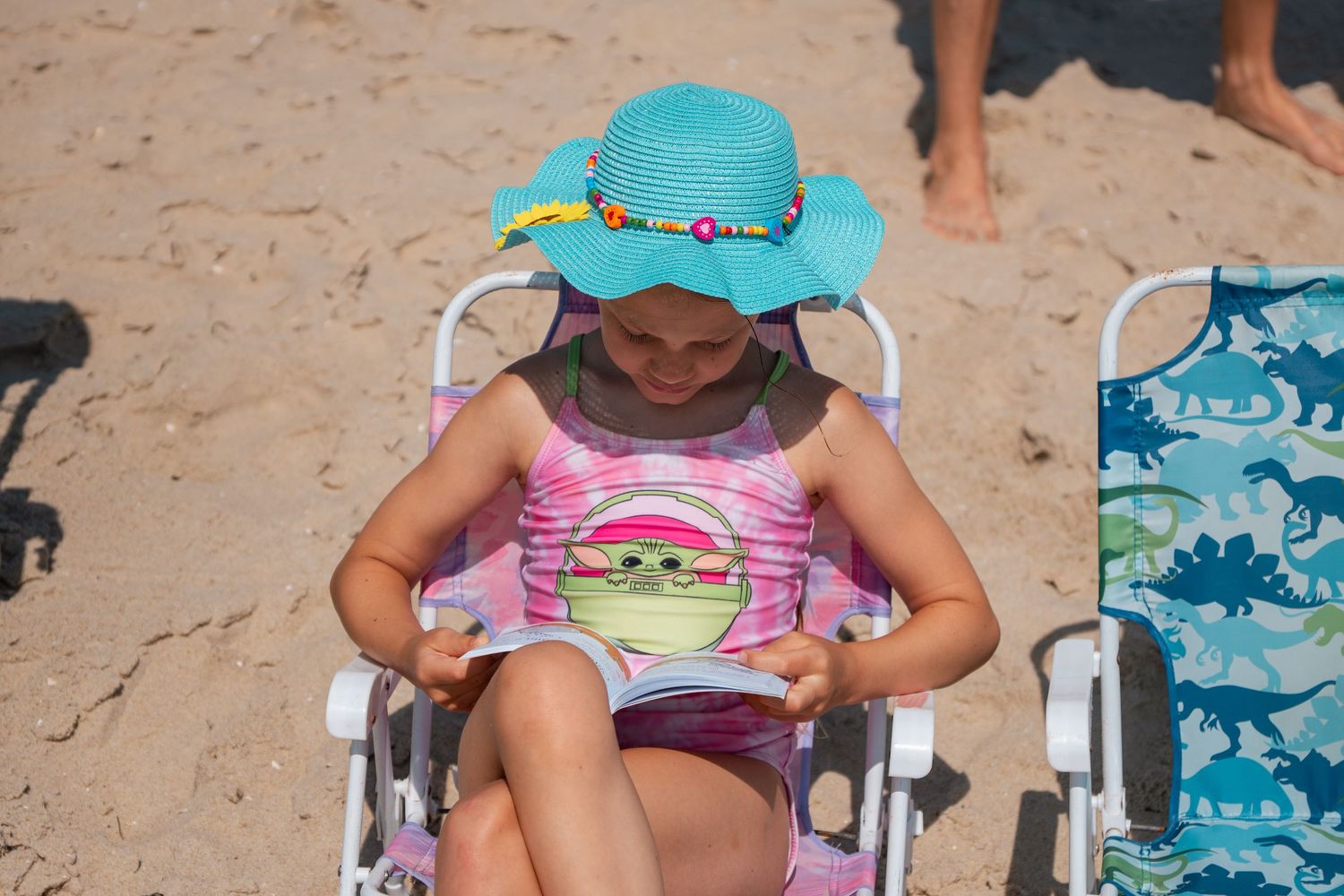 Young girl in teal sun hat and pink swimsuit reading on beach chair at sandy beach.