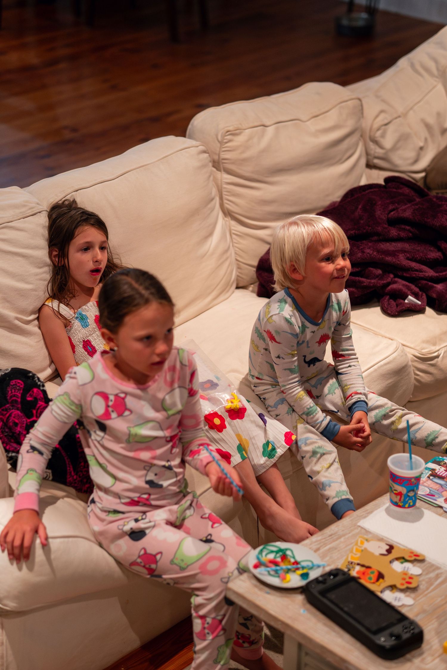 Three children in pajamas sitting on a white sofa with snacks on a coffee table.