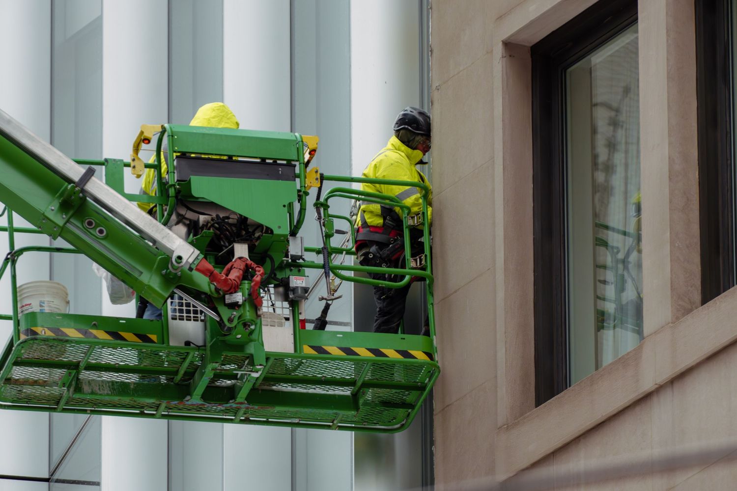 Two workers in high-visibility jackets operating green aerial work platform on building.