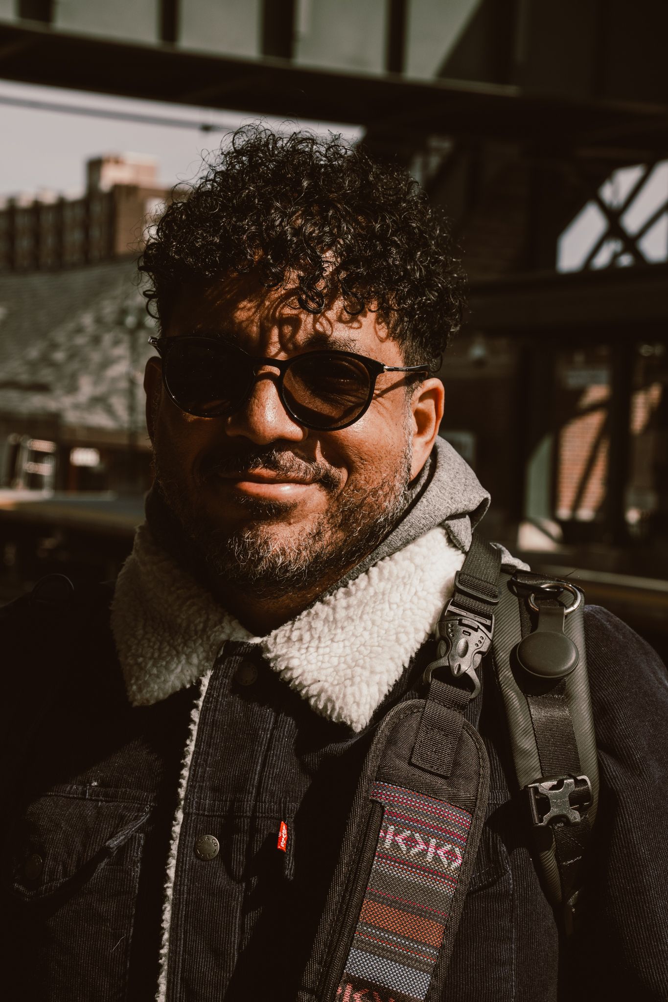 Portrait of a curly-haired bearded man in sunglasses and sherpa jacket with a camera strap at a train station.