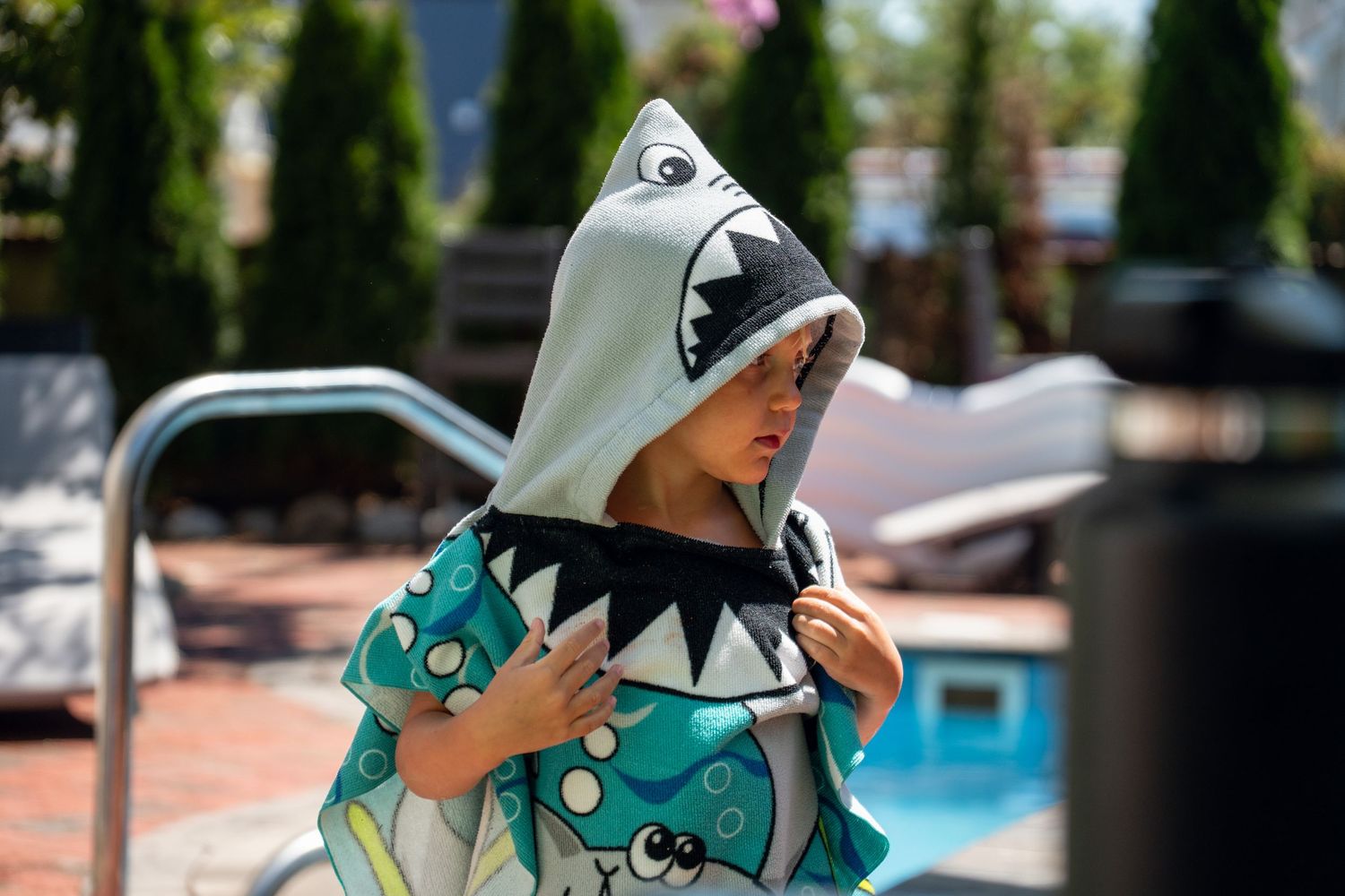 Young child wearing a shark hooded towel poncho standing by a backyard swimming pool.