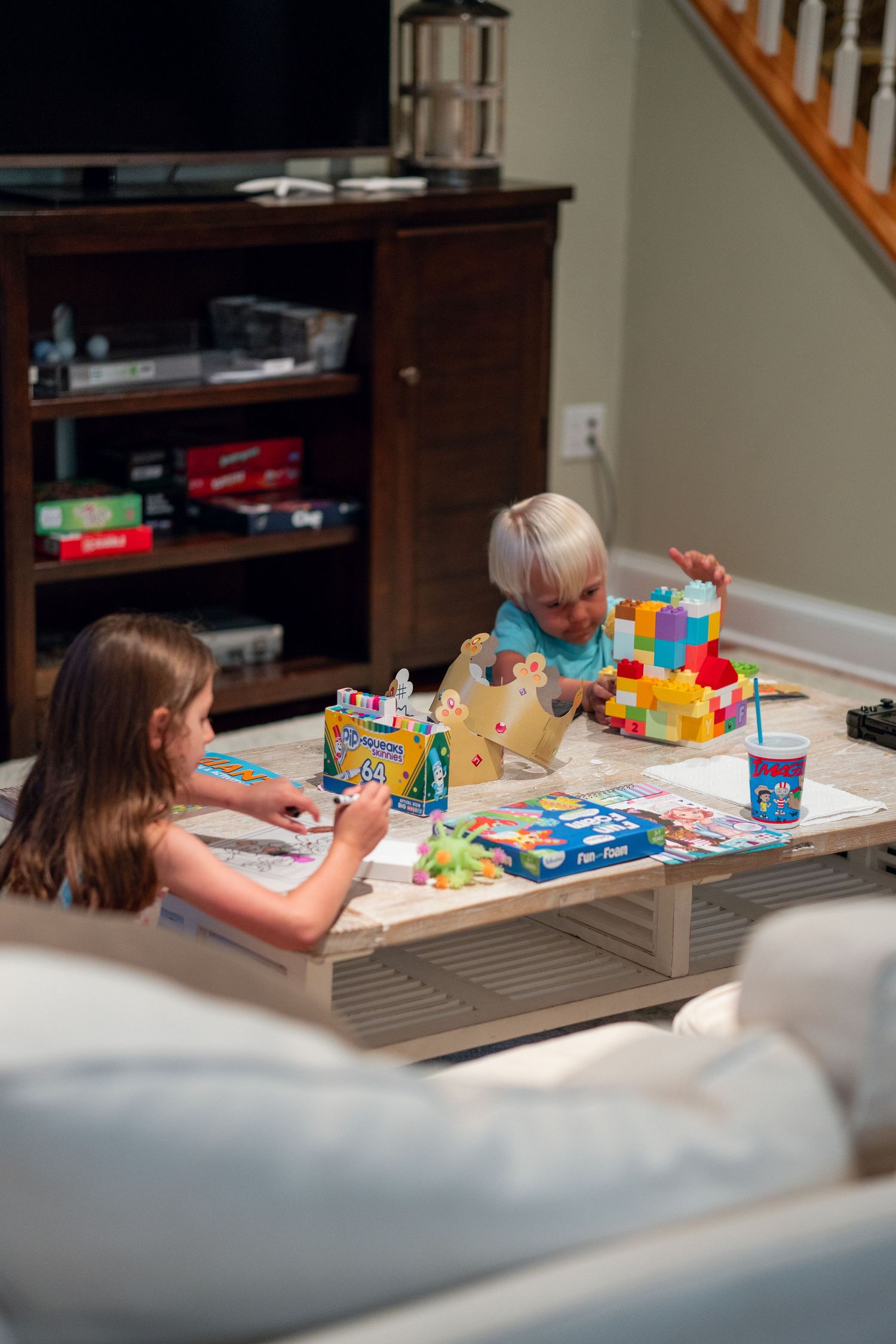 Two young children playing with colorful building blocks and art supplies on a coffee table indoors.