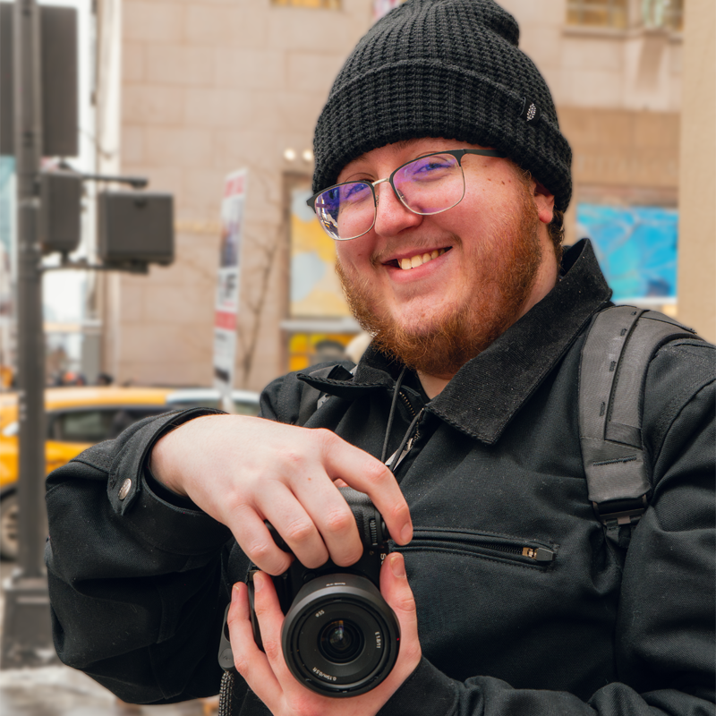 A smiling bearded man wearing a black beanie and glasses holding a DSLR camera outdoors.