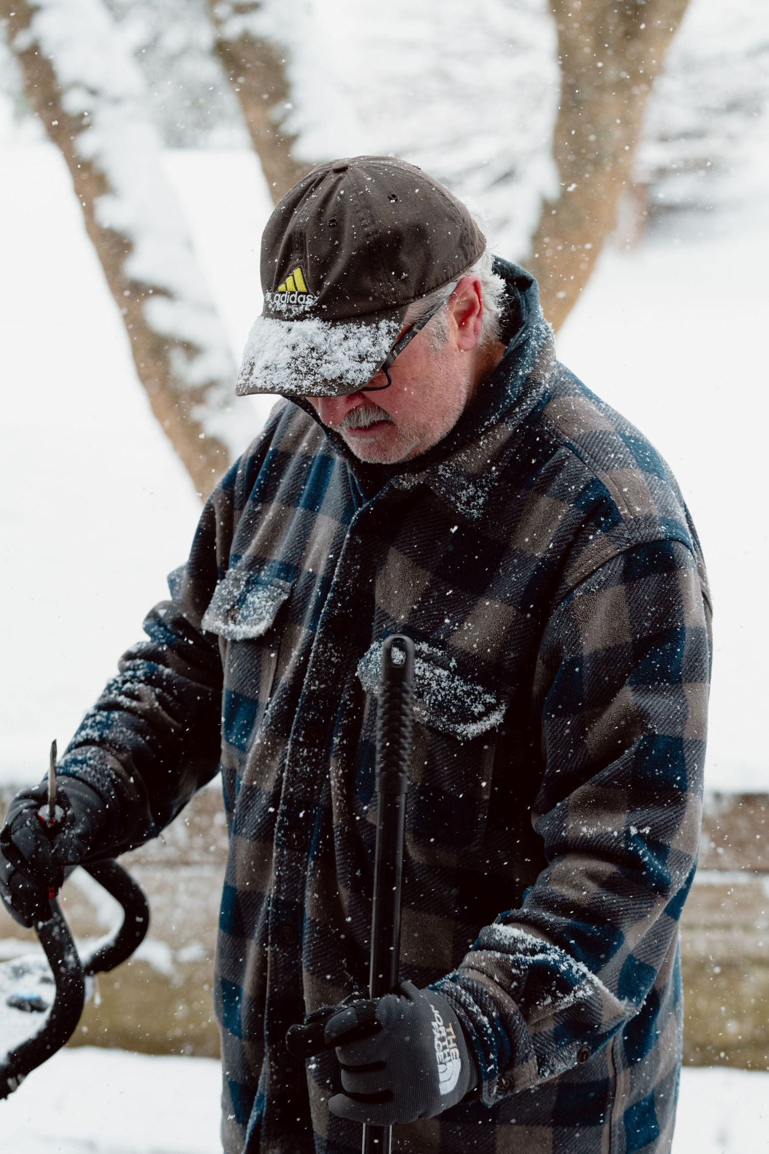 Older man in plaid jacket adjusting snowblower controls while snow falls around him outdoors.