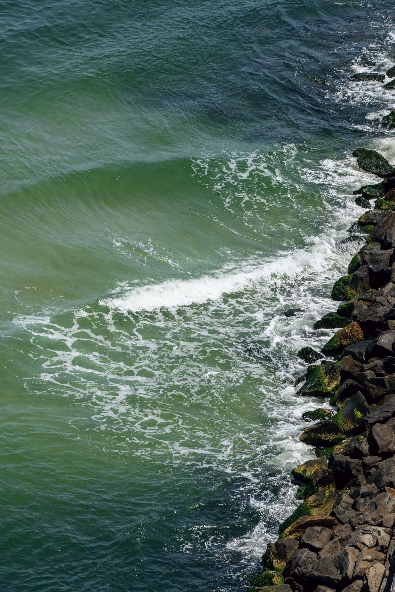 Aerial view of green ocean waves crashing against a rocky shoreline with white sea foam and dark mossy stones.