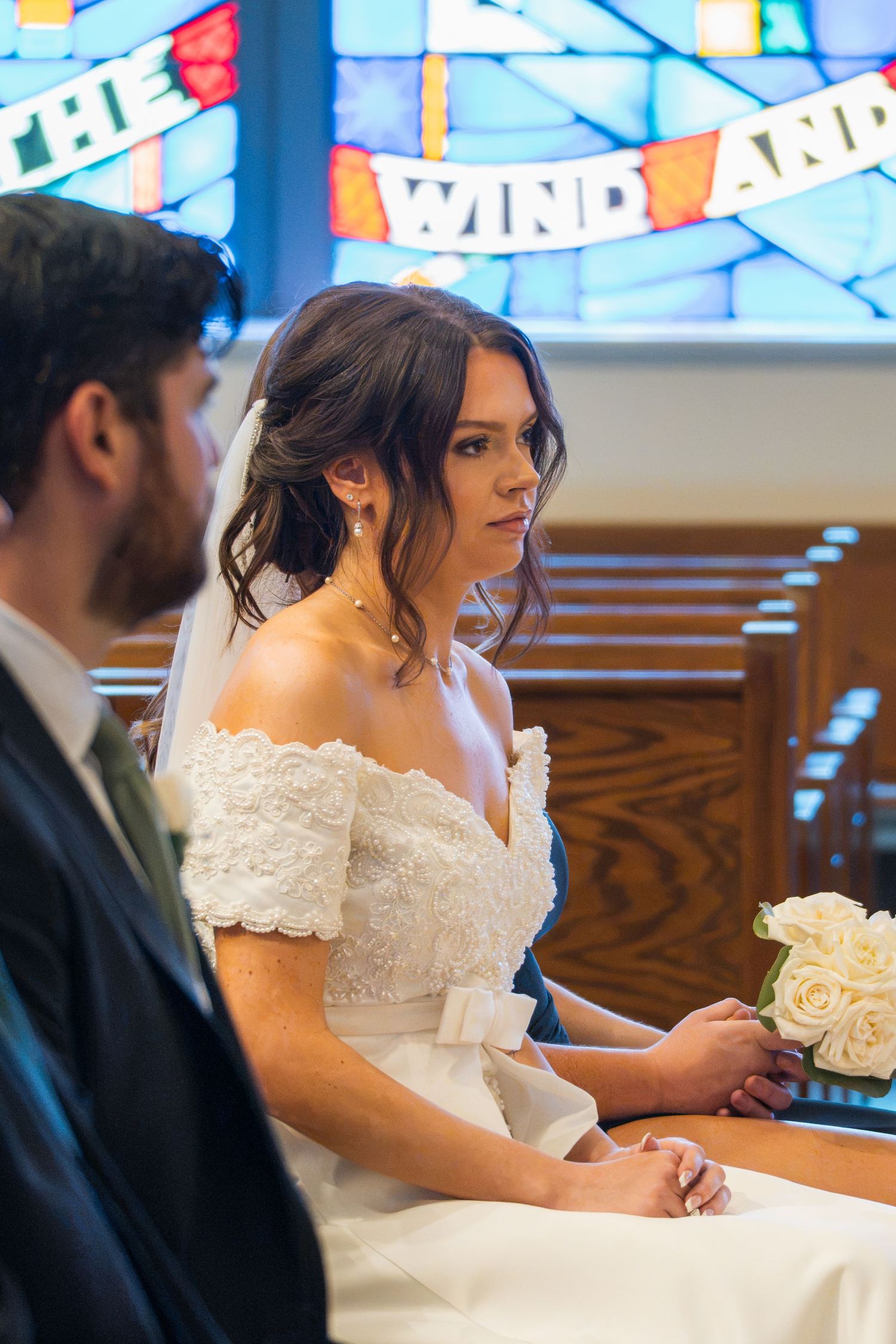 Bride in white off-shoulder lace gown holding roses during church wedding ceremony with stained glass window backdrop.
