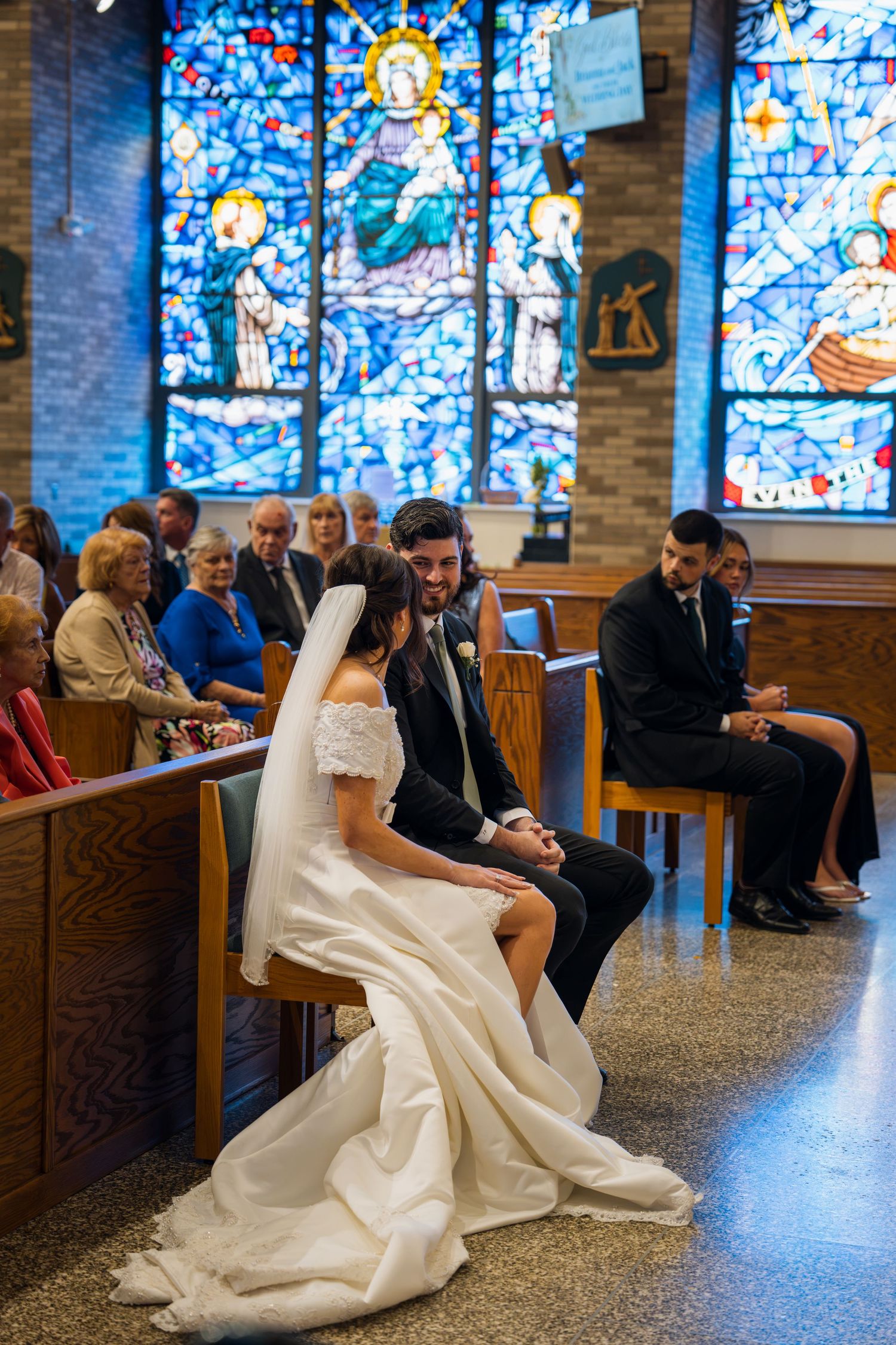 Bride and groom sit together in church pew during wedding ceremony with colorful stained glass windows behind them.