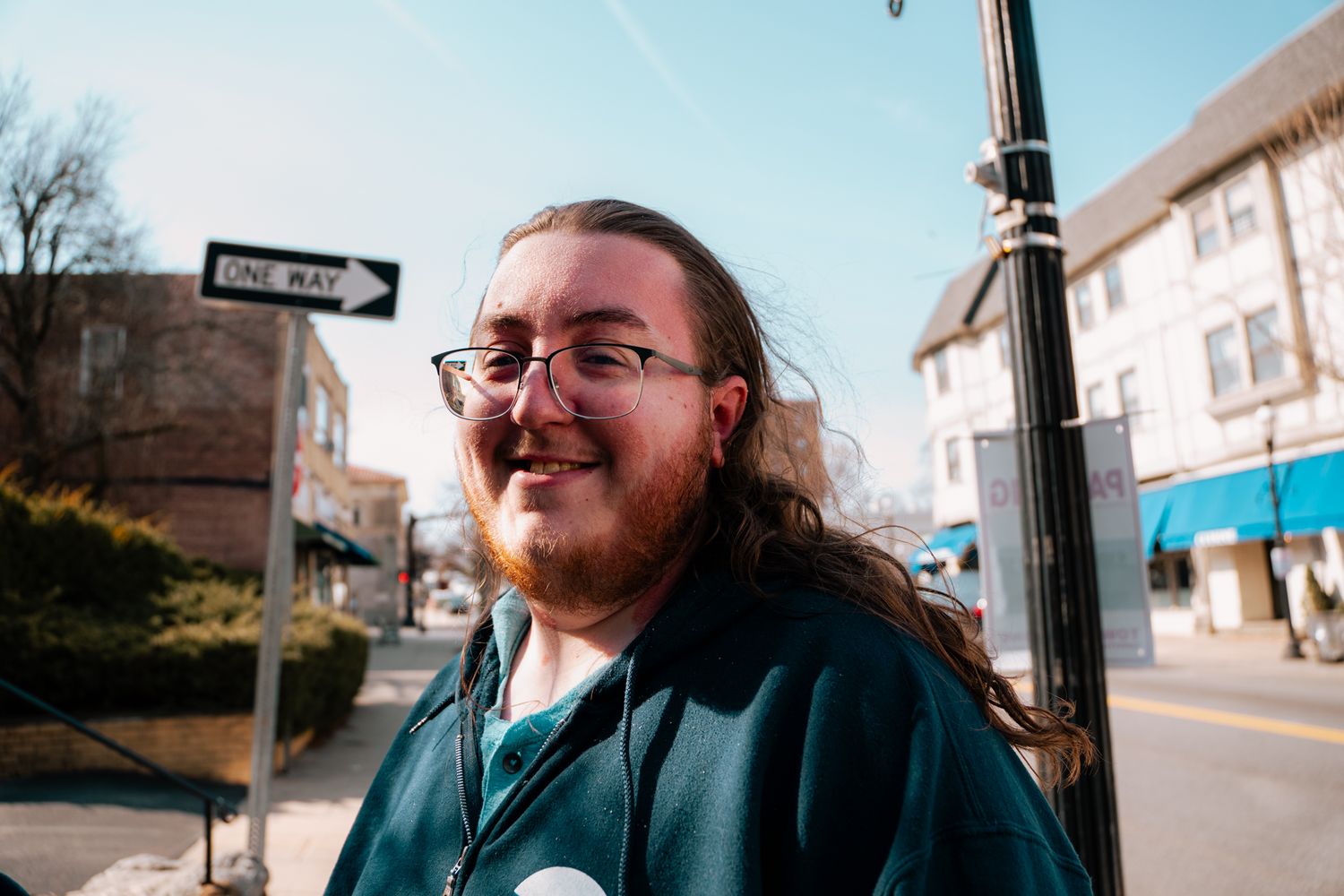Smiling young man with long hair and glasses standing on a sunny urban street.
