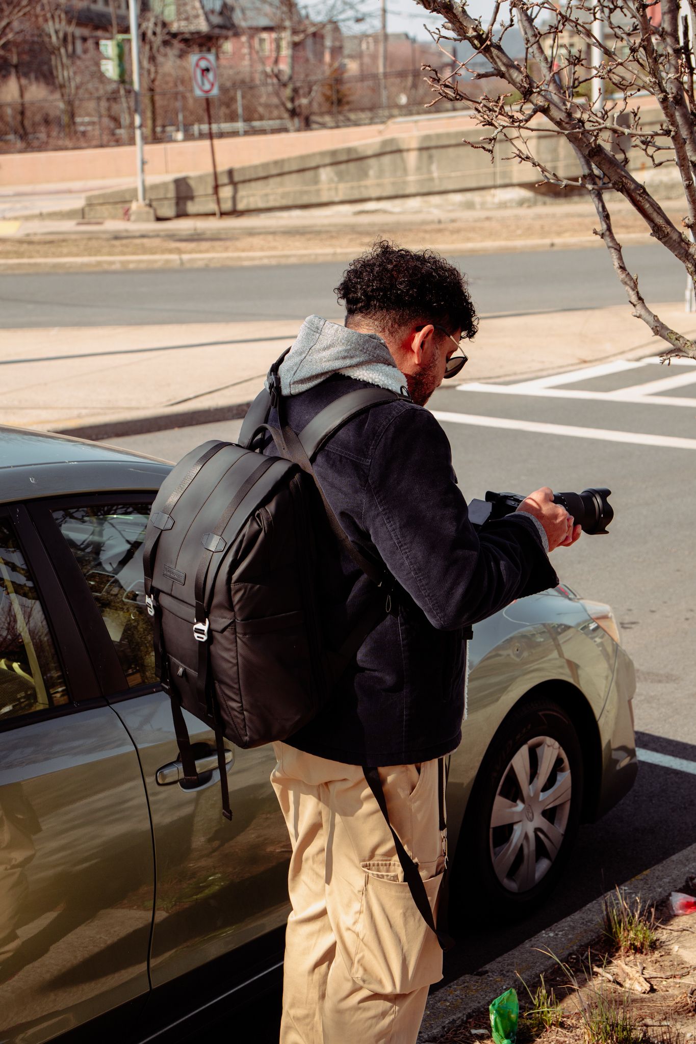 A curly-haired photographer with a backpack and camera stands beside a car on a sunny urban street.