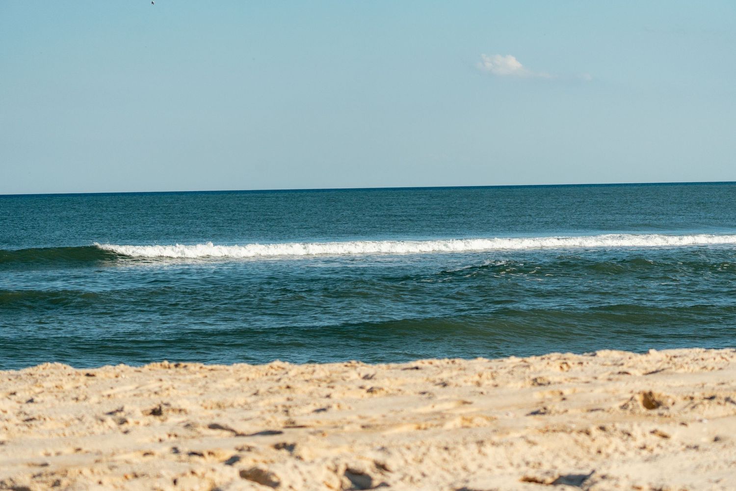 Gentle ocean waves break on a sandy beach shoreline under a bright blue sky on a clear sunny day.