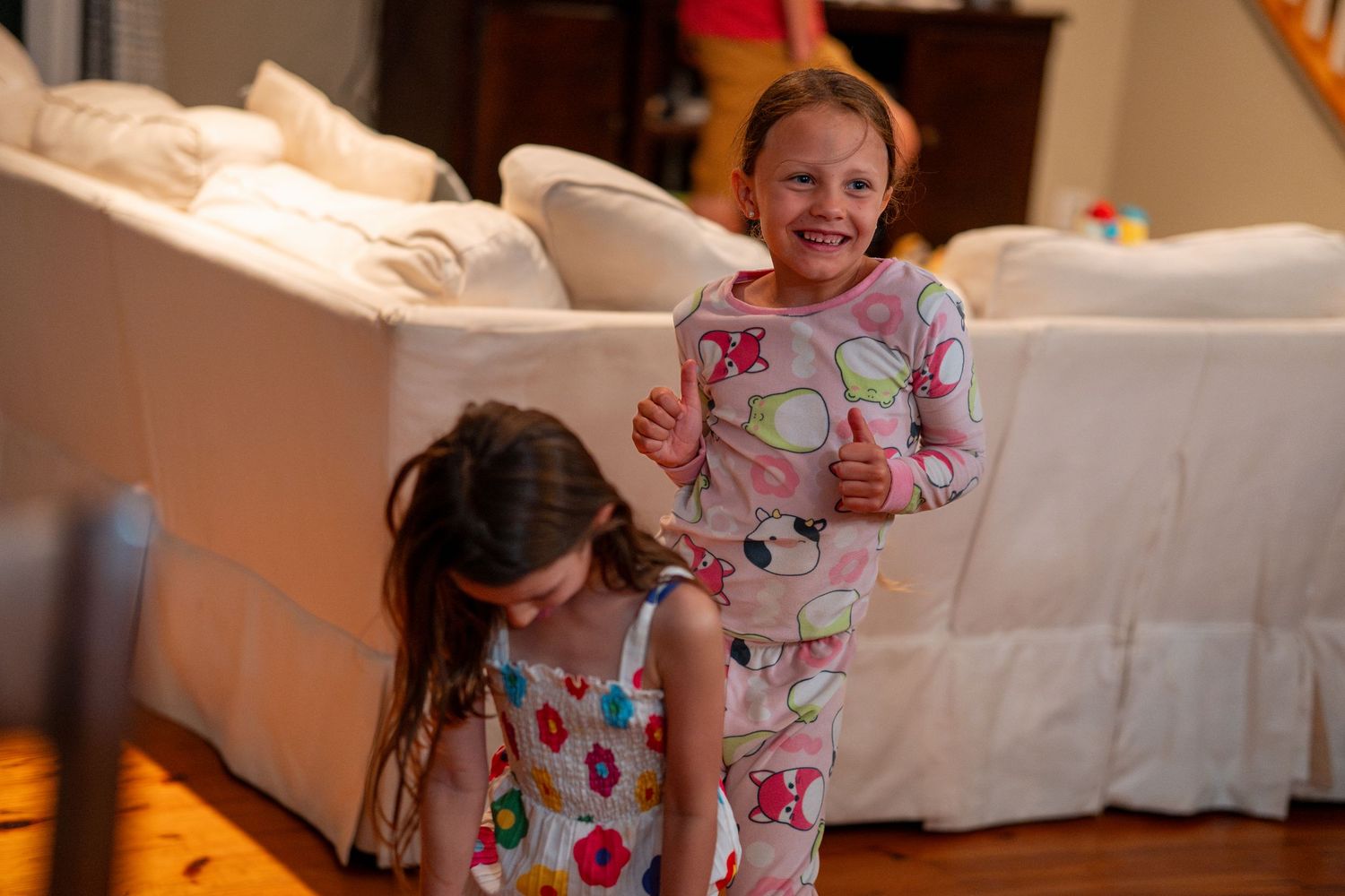 Two young girls in pajamas smile and play together in a warmly lit living room.