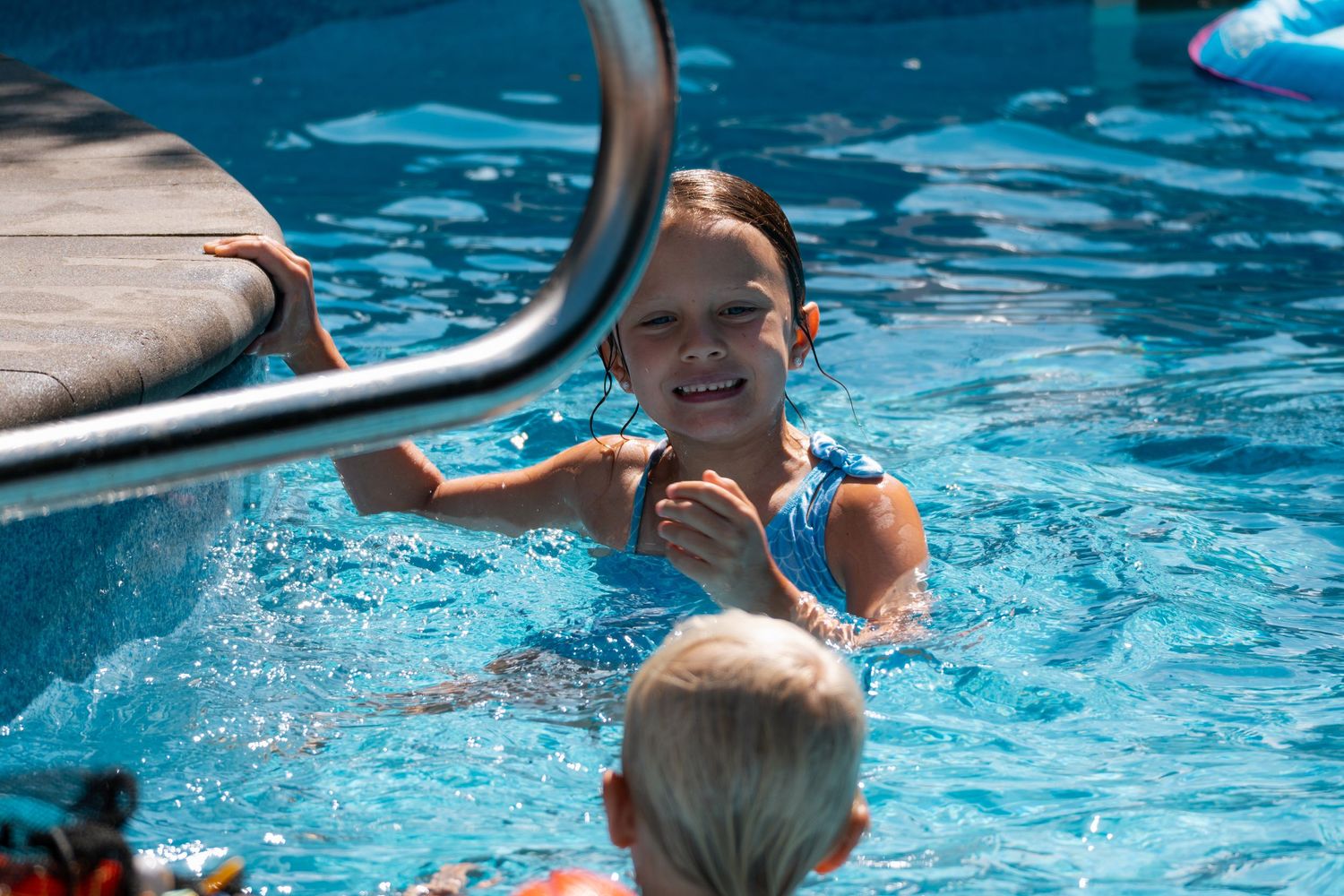 Smiling girl in blue swimsuit enjoying a sunny outdoor swimming pool near the ladder.