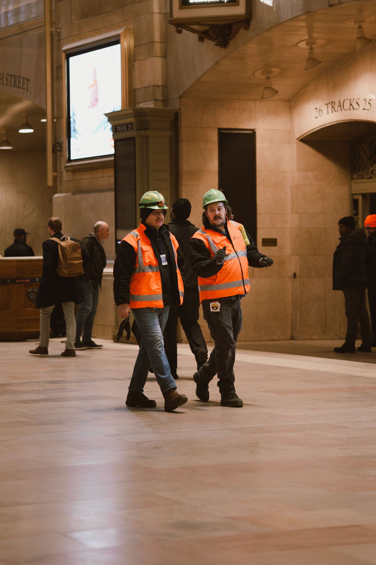 Two construction workers in orange vests and green helmets walk through Grand Central Terminal.