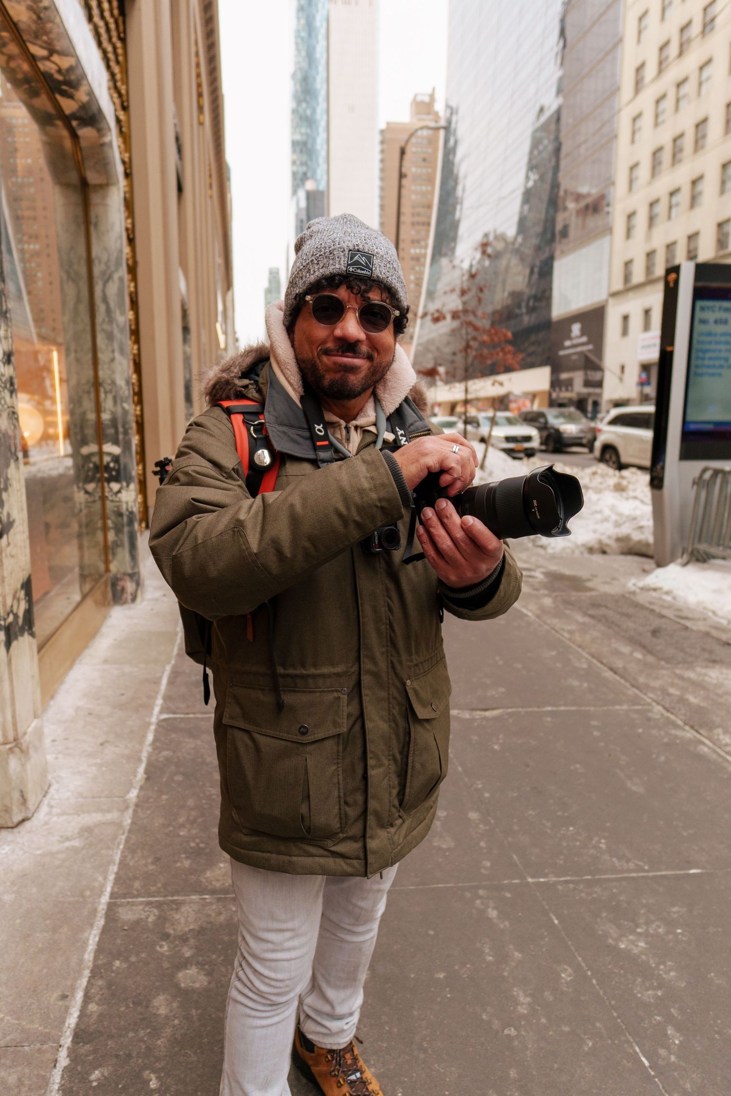 A photographer in a green parka and gray beanie holds a camera on a snowy New York City sidewalk.