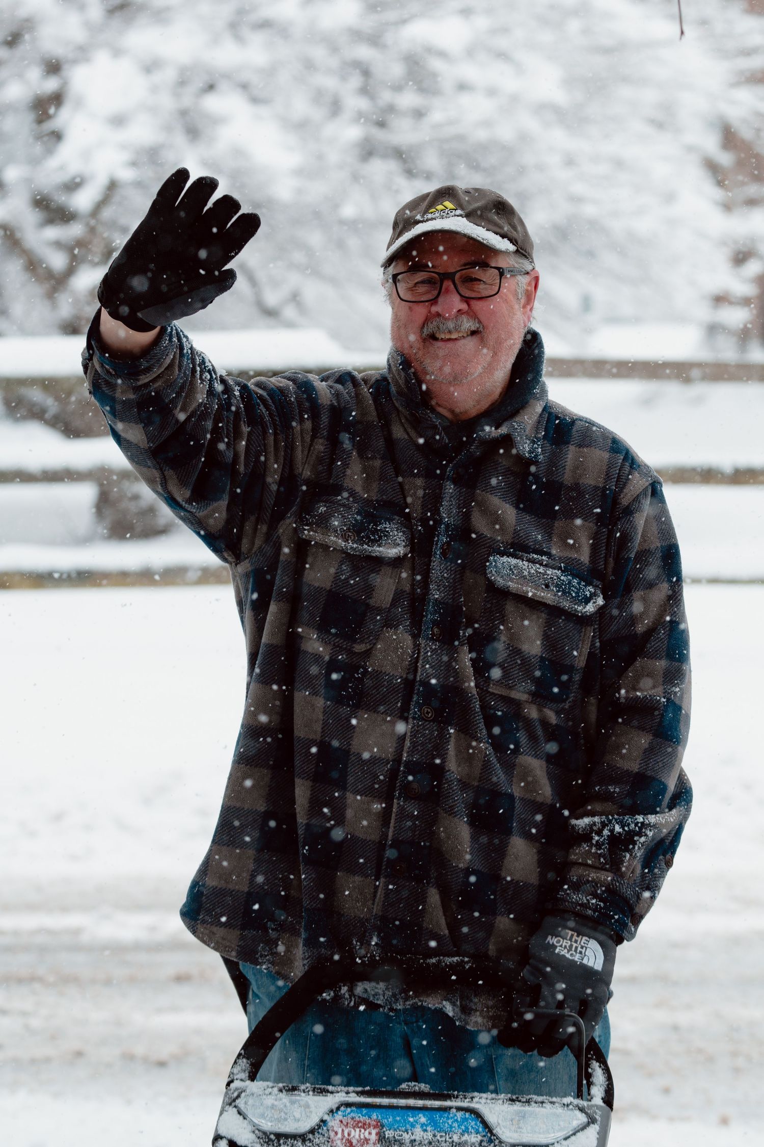 Smiling elderly man in plaid jacket waving while using a snowblower during a snowstorm.