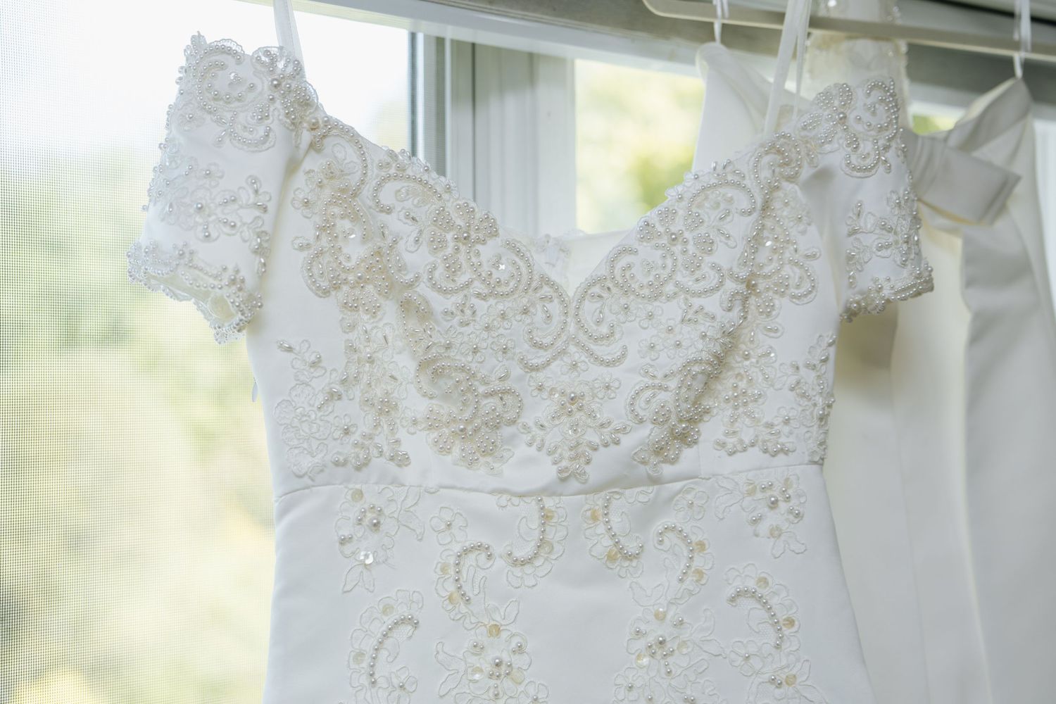 White beaded lace wedding dress hanging on a hanger near a bright window, featuring intricate embroidery details.