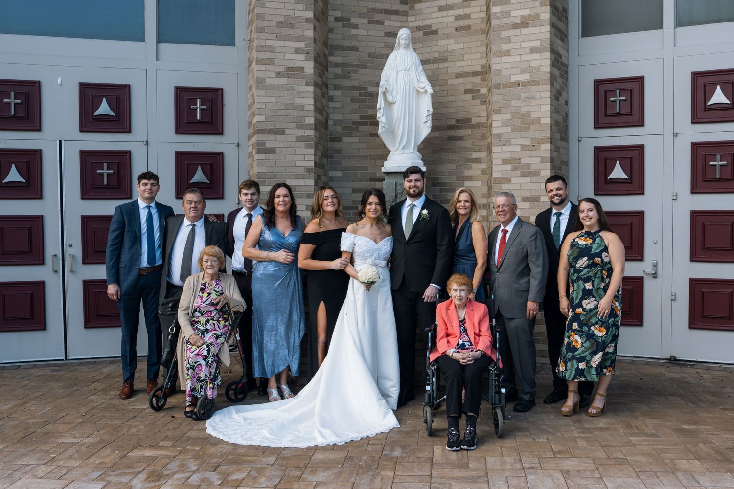 Wedding party poses in front of a church statue of Mary, bride in white gown with family in formal attire.