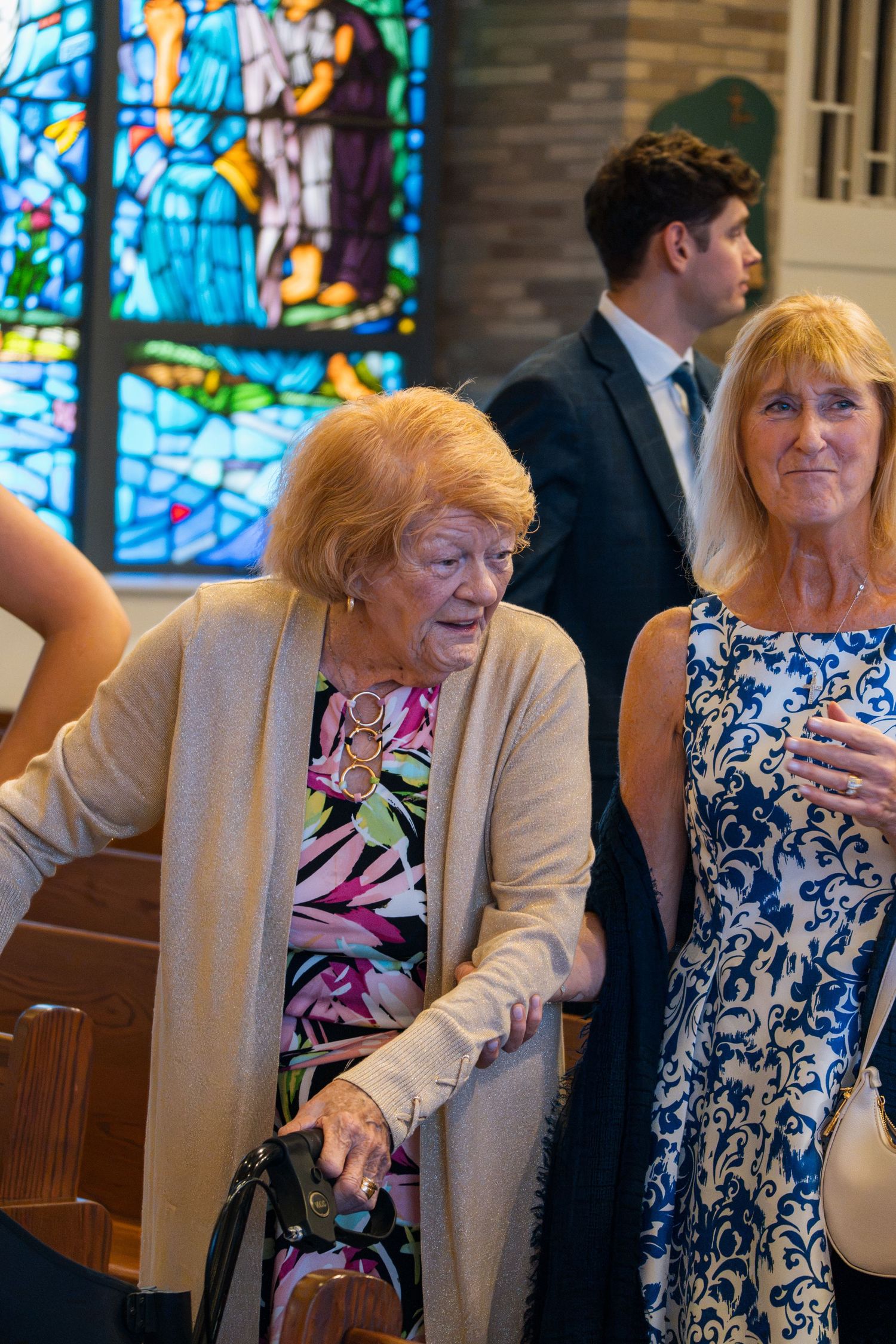 Two women walking through a church with stained glass windows during a formal event.
