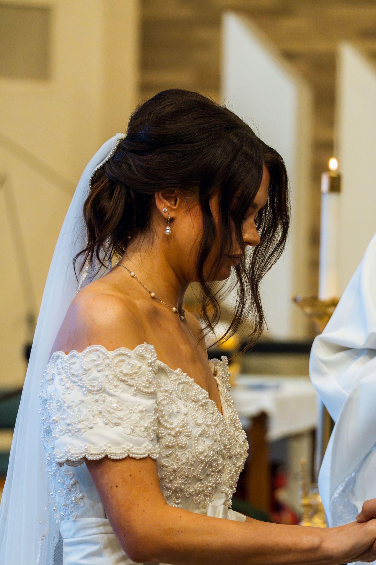 Bride in lace off-shoulder wedding dress with veil, bowing head during church ceremony near candlelit altar.