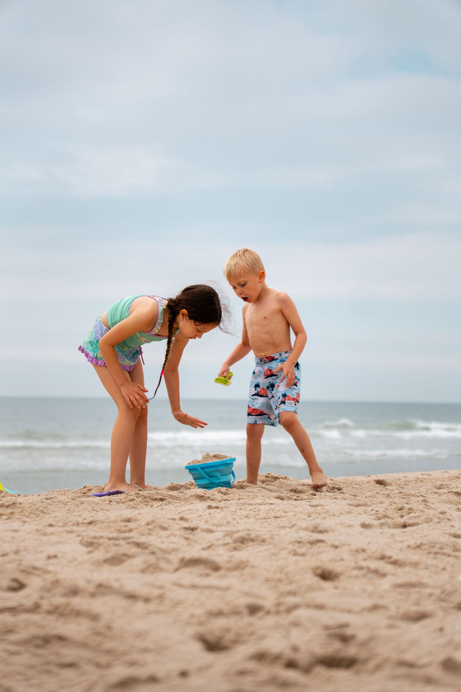 Two children playing on a sandy beach near the ocean, with a girl in a swimsuit and a boy in swim trunks.
