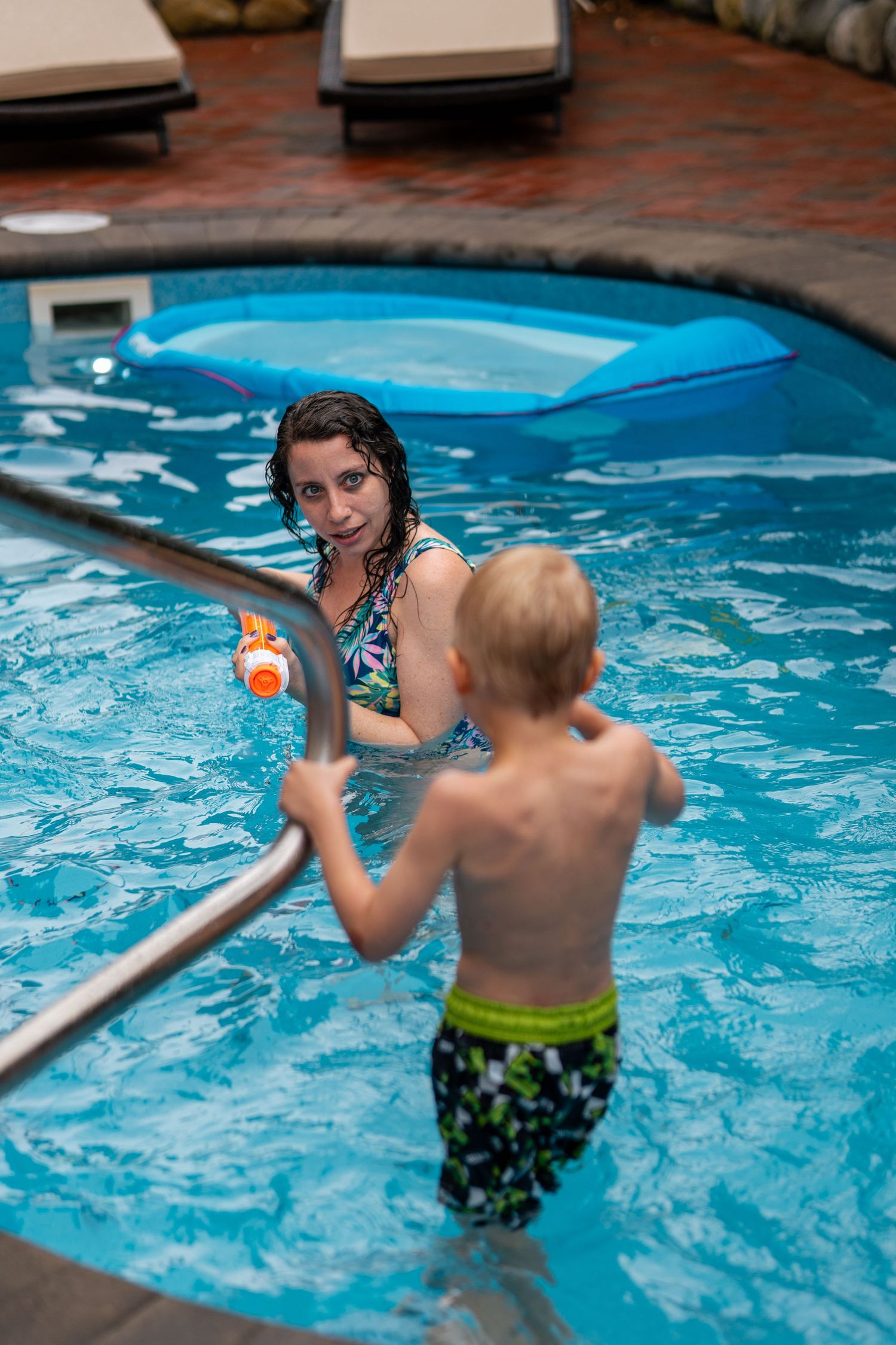 A woman and young boy playing with a water gun in an indoor swimming pool.