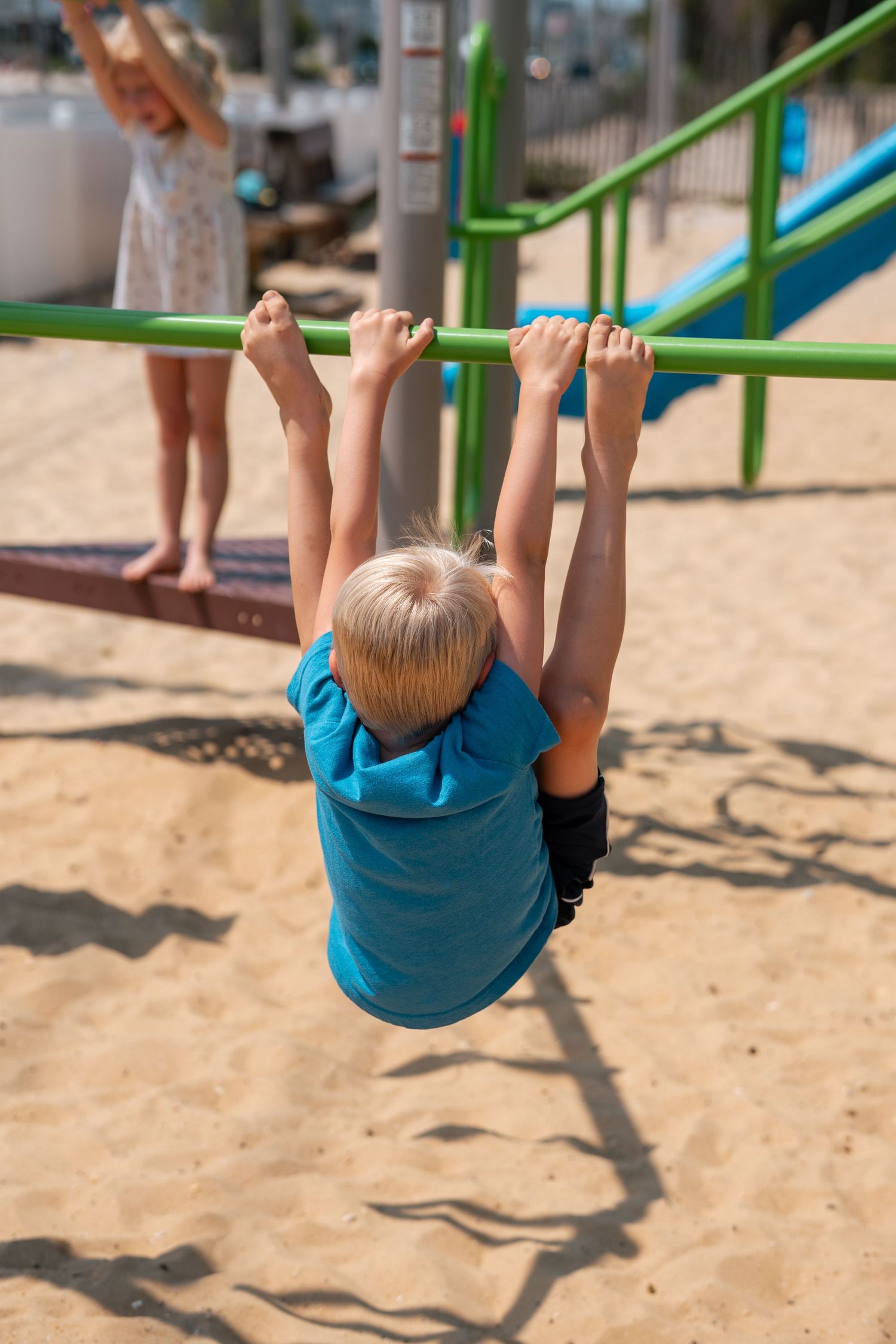 Blonde child hanging from green monkey bars on sandy playground while another child plays in the background.