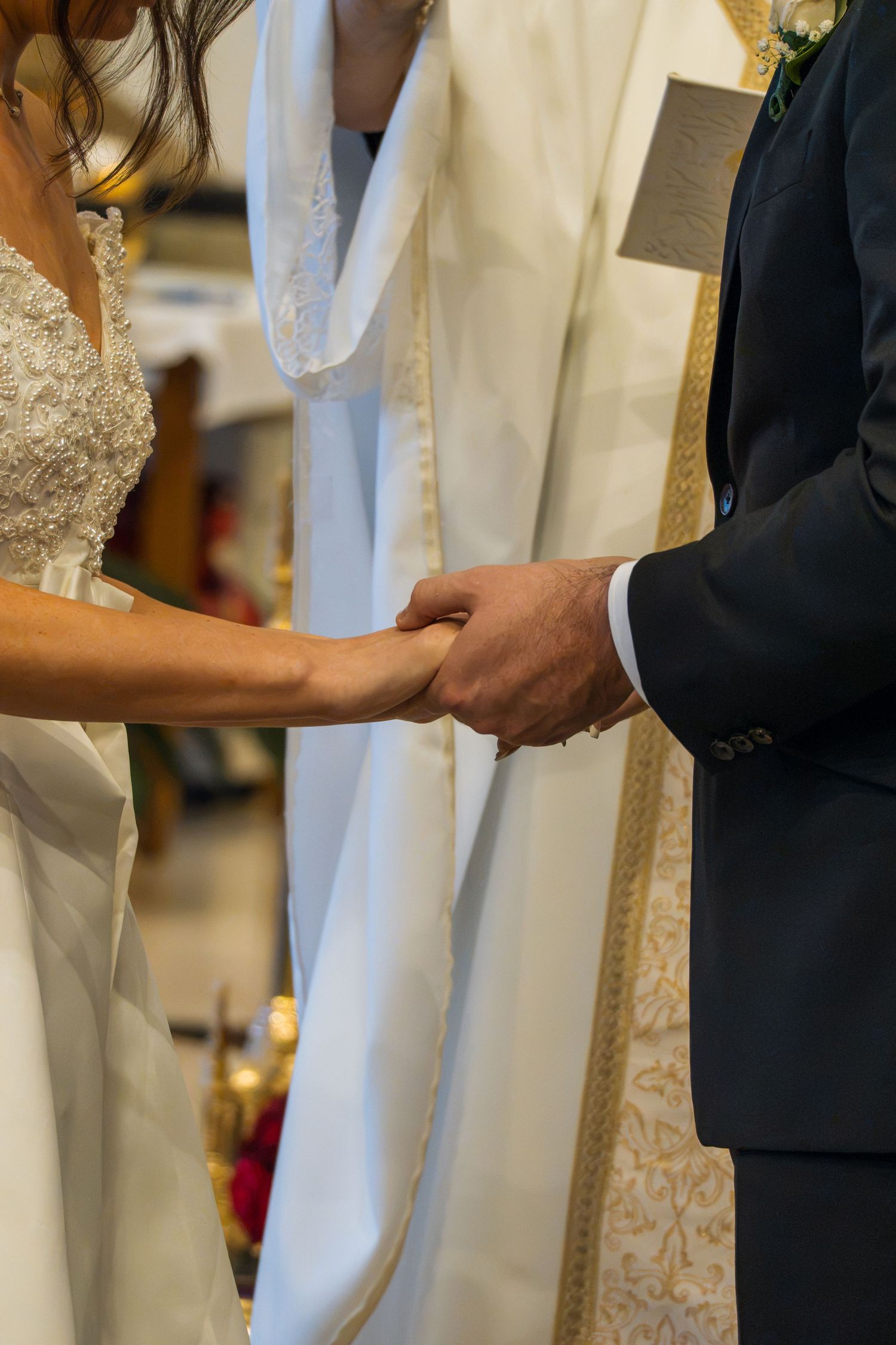 Bride and groom holding hands during wedding ceremony, bride in lace gown, groom in navy suit, priest in gold vestments.