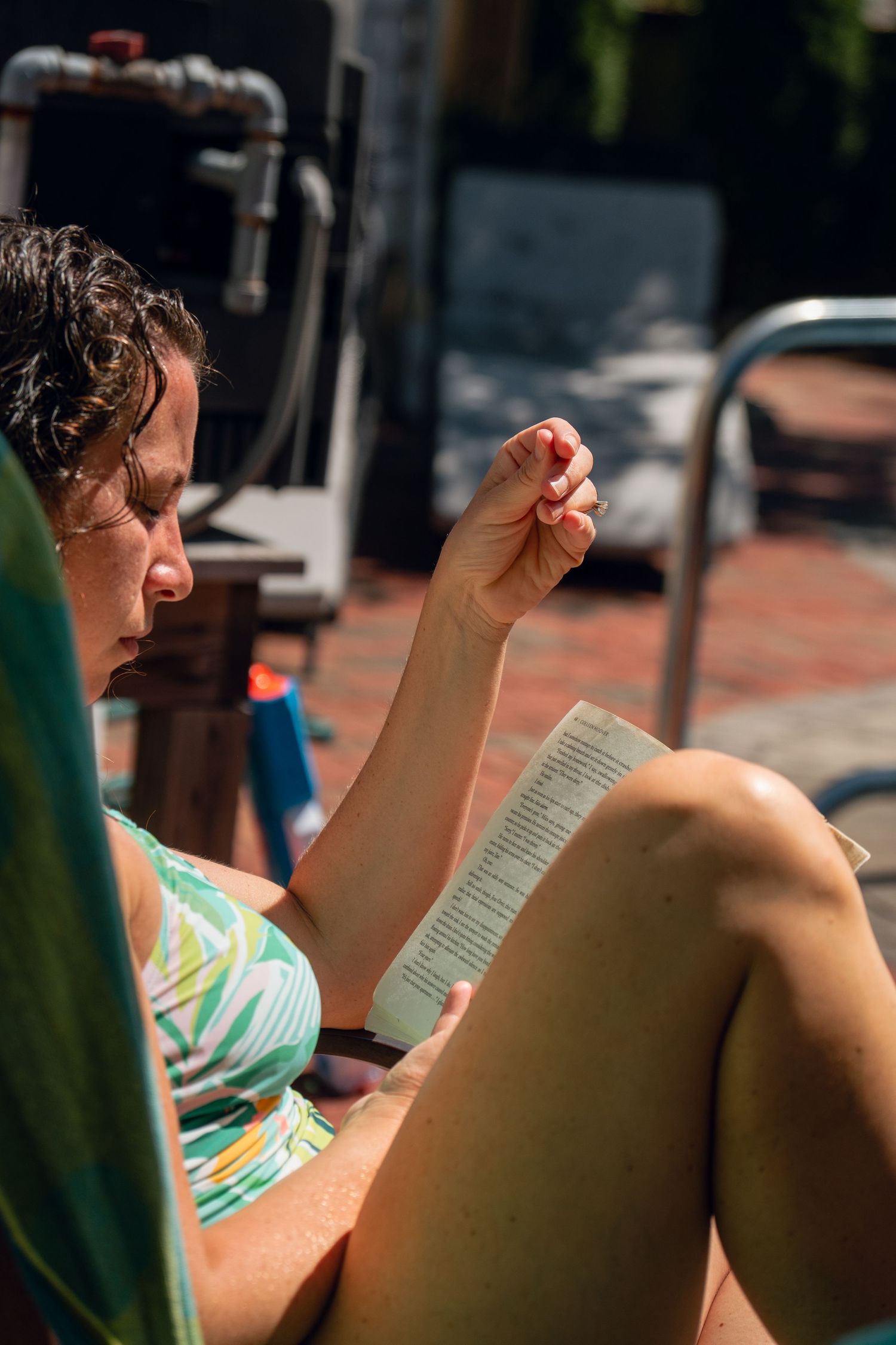 Woman in bikini reading a book poolside on a sunny day, raising her hand while lounging.