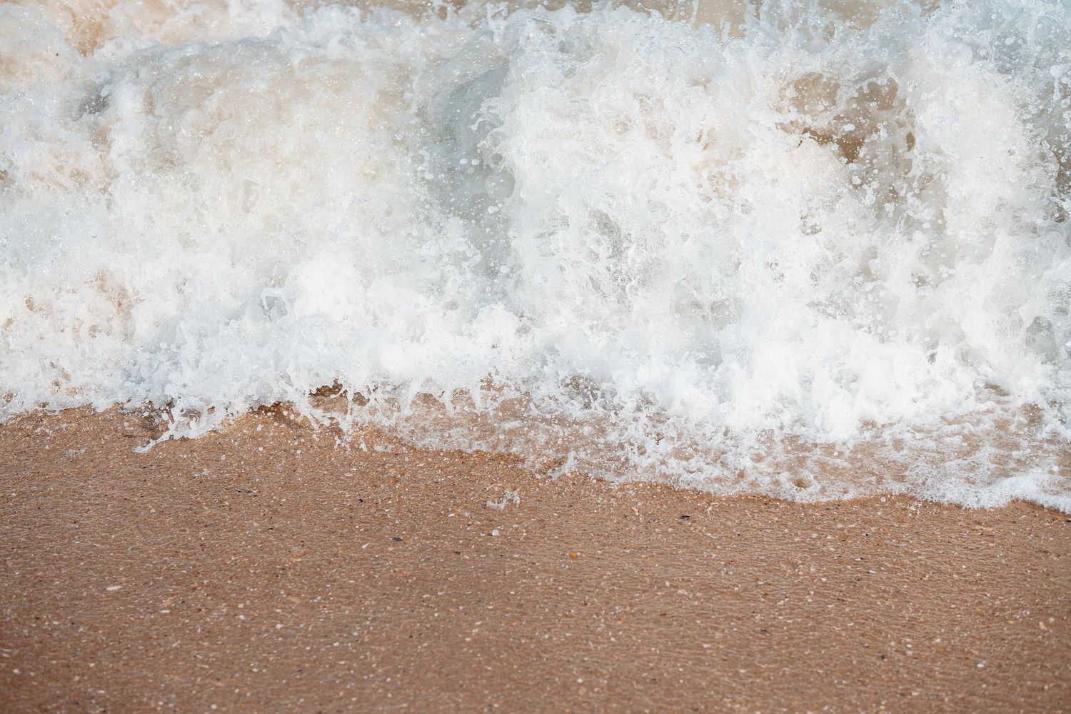 White sea foam from ocean wave spreading across wet sandy beach shoreline close-up.